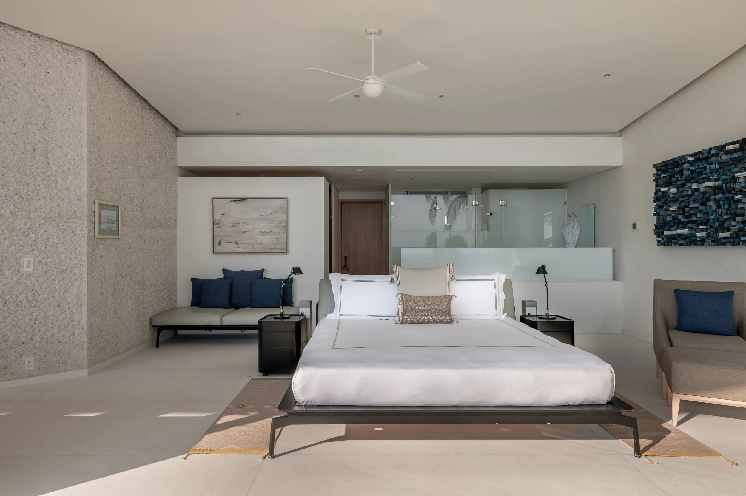 Modern minimalist hotel room with white bed, navy pillows, black bedside tables with lamps, a framed abstract painting, textured wall, beige armchair, and large glass window with palm trees outside.