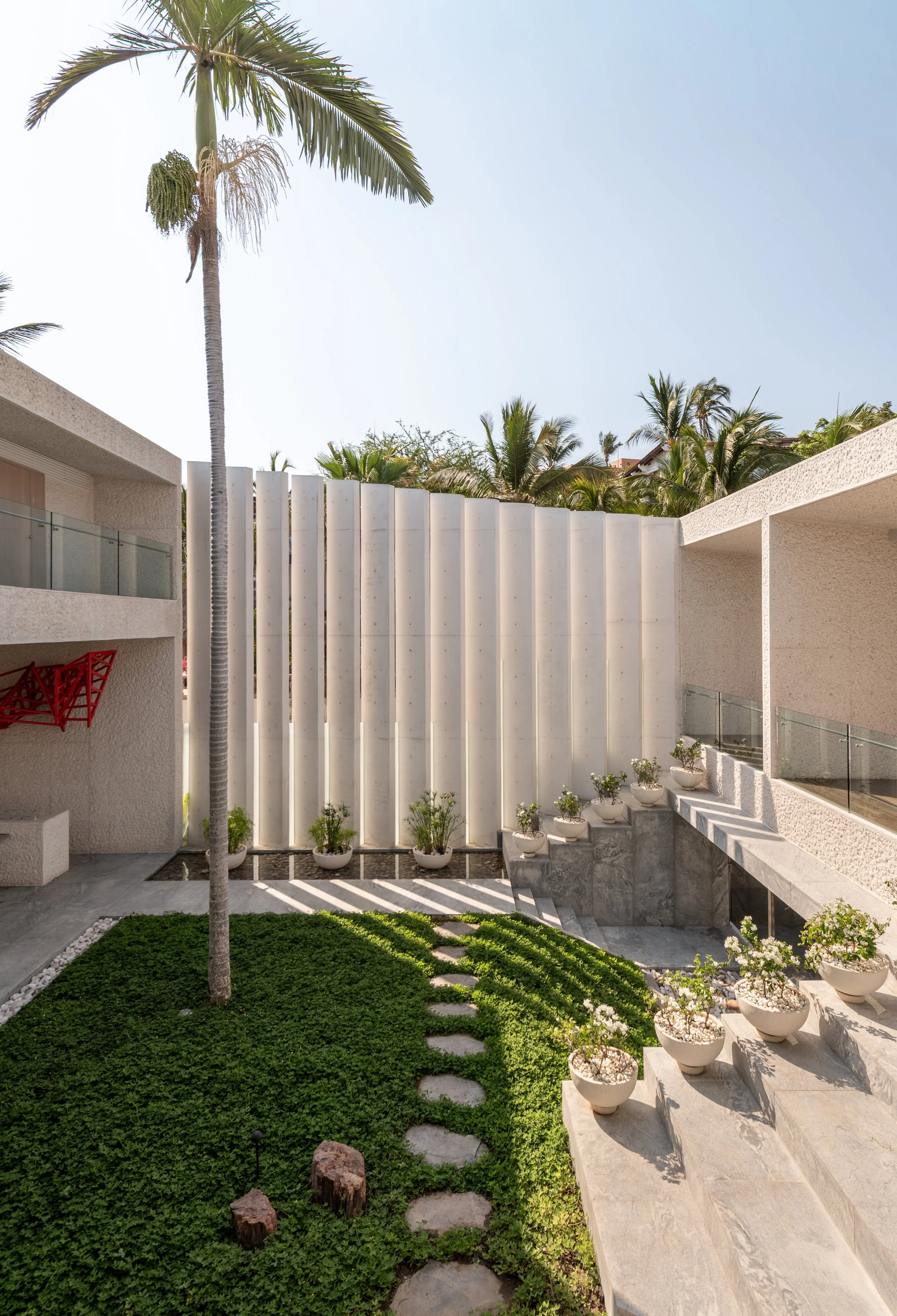 A modern courtyard with a small green lawn, potted plants lining the steps, a tall palm tree, and a white wall with trees and a clear blue sky in the background.