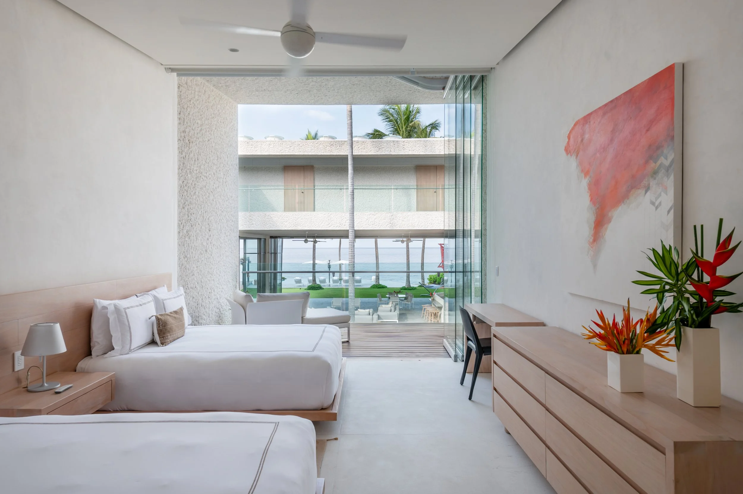 View of a luxurious hotel room with white beds, a wooden dresser with a potted plant, and a balcony overlooking palm trees, ocean, and blue sky.
