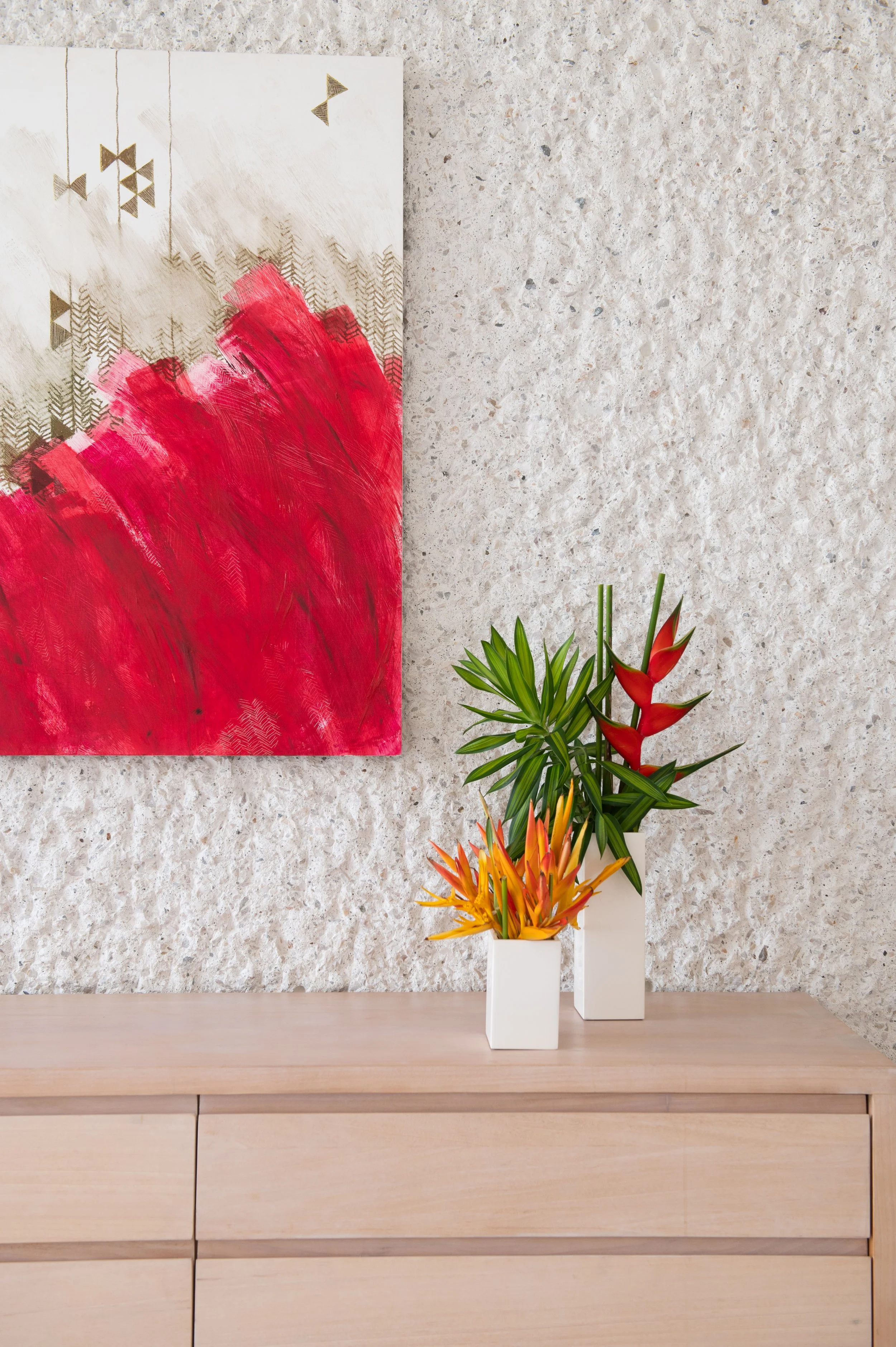A modern interior with a textured wall, abstract art featuring red and gold designs, and two white vases with green and red tropical flowers on a wooden cabinet.