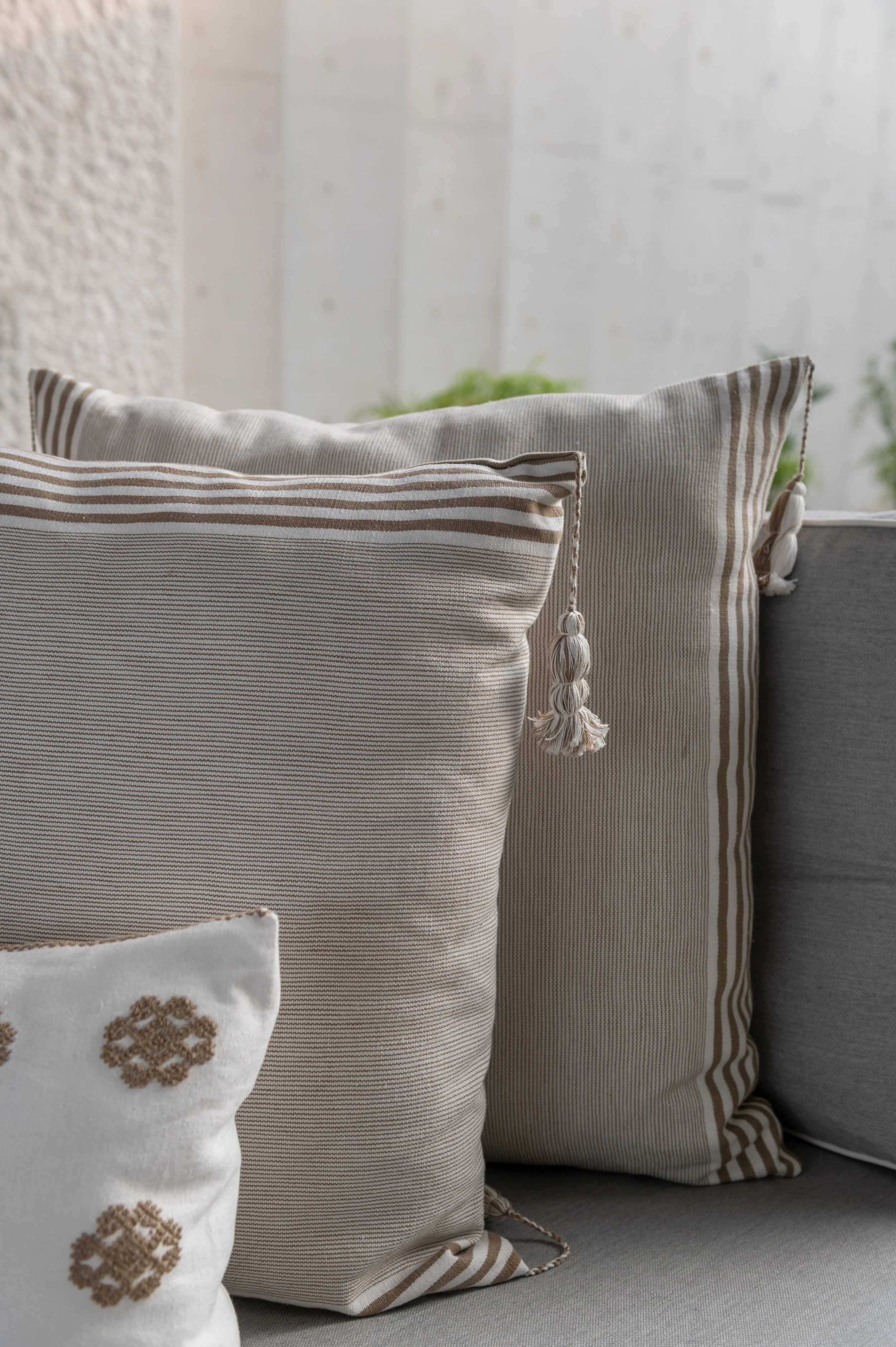 A collection of neutral-colored throw pillows with striped and embroidered patterns arranged on a light gray sofa, with a textured wall and green plants in the background.