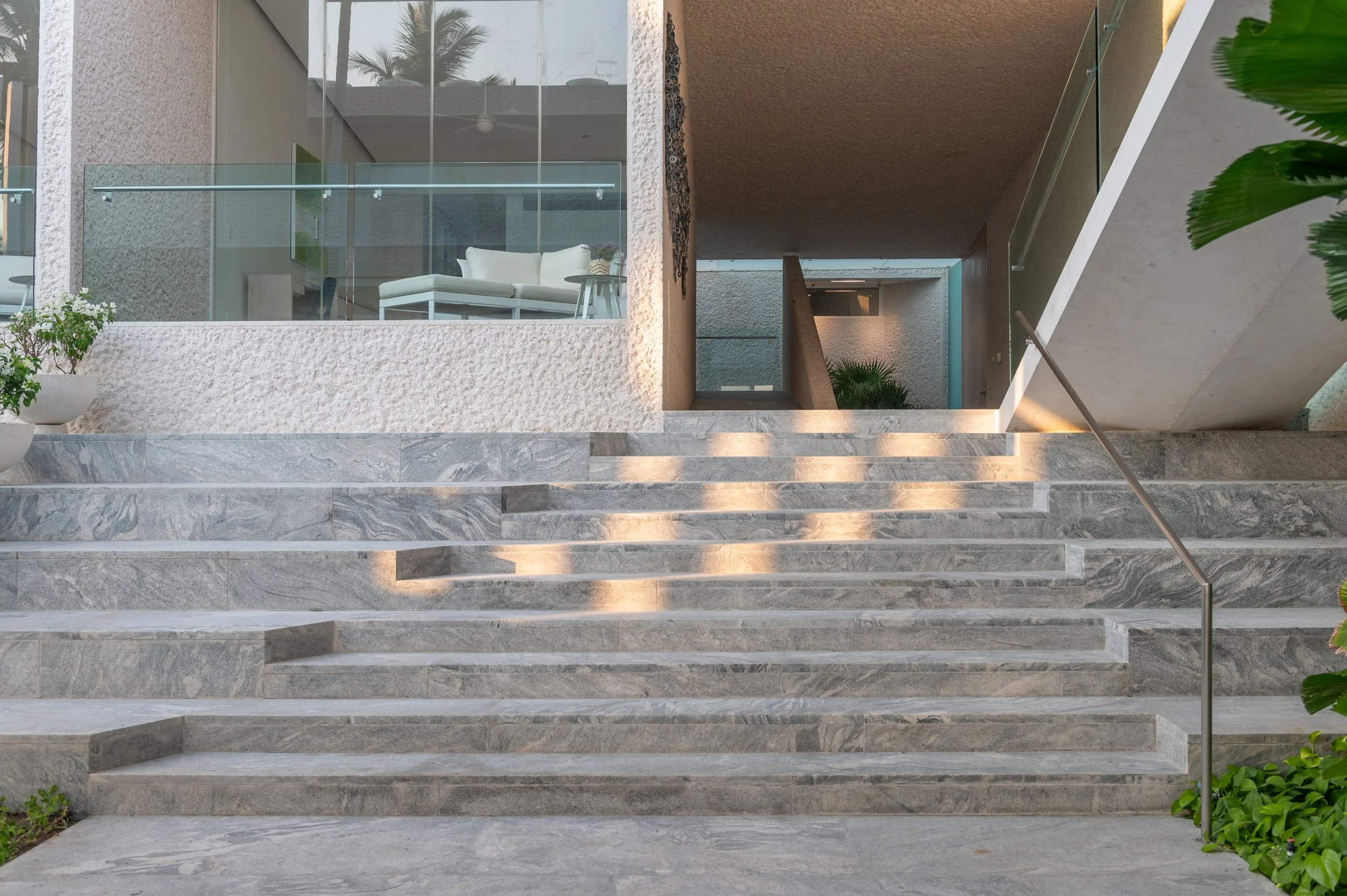 Modern building with stone stairs leading up to an entrance with glass railings and a seating area visible through a large window, surrounded by palm trees and greenery.