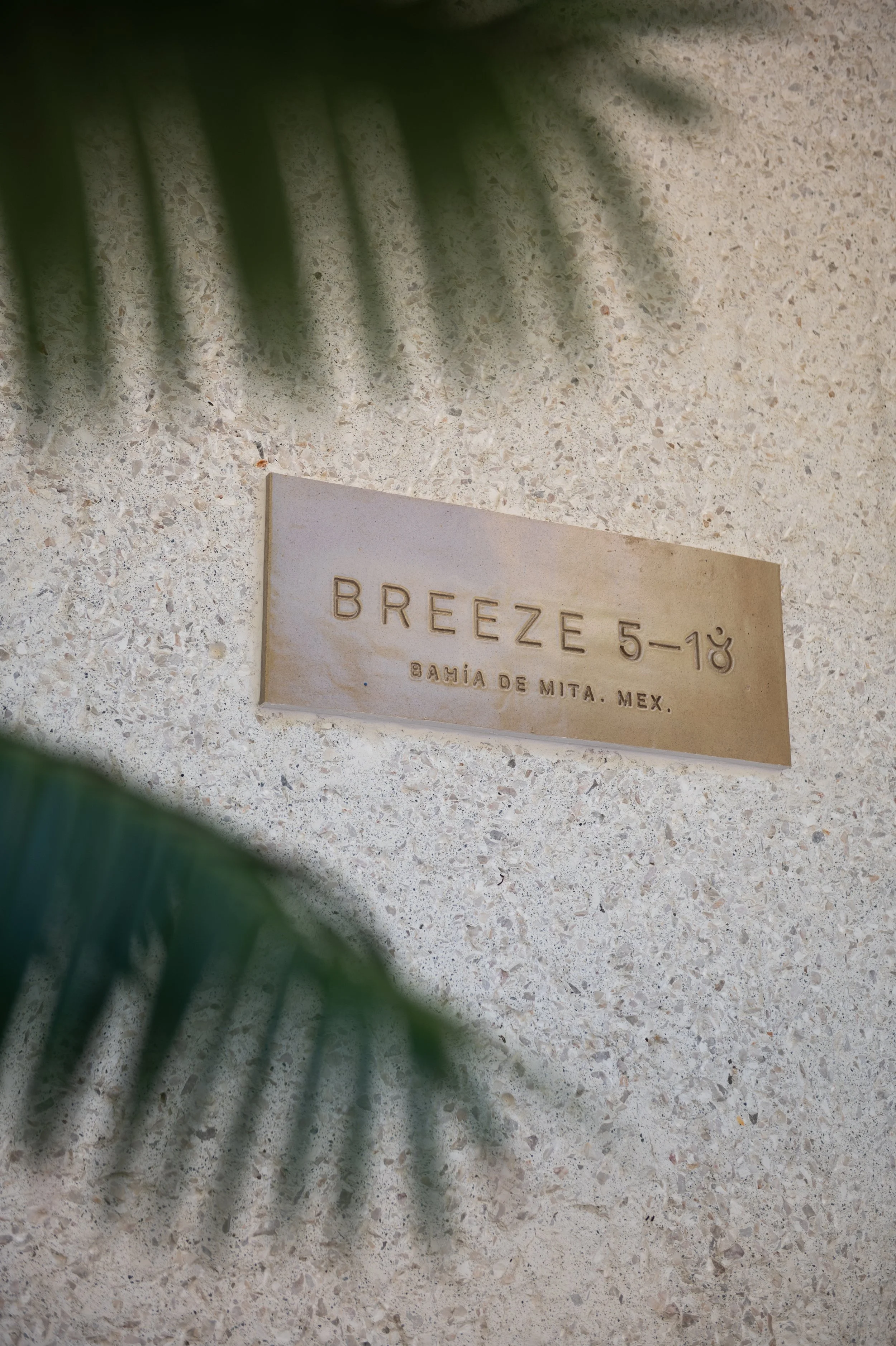 Close-up of a beige terrazzo wall with a beige plaque labeled 'BREEZE 5-10' and 'BAHIA DE MITA, MEX.' partially obscured by green plant leaves.