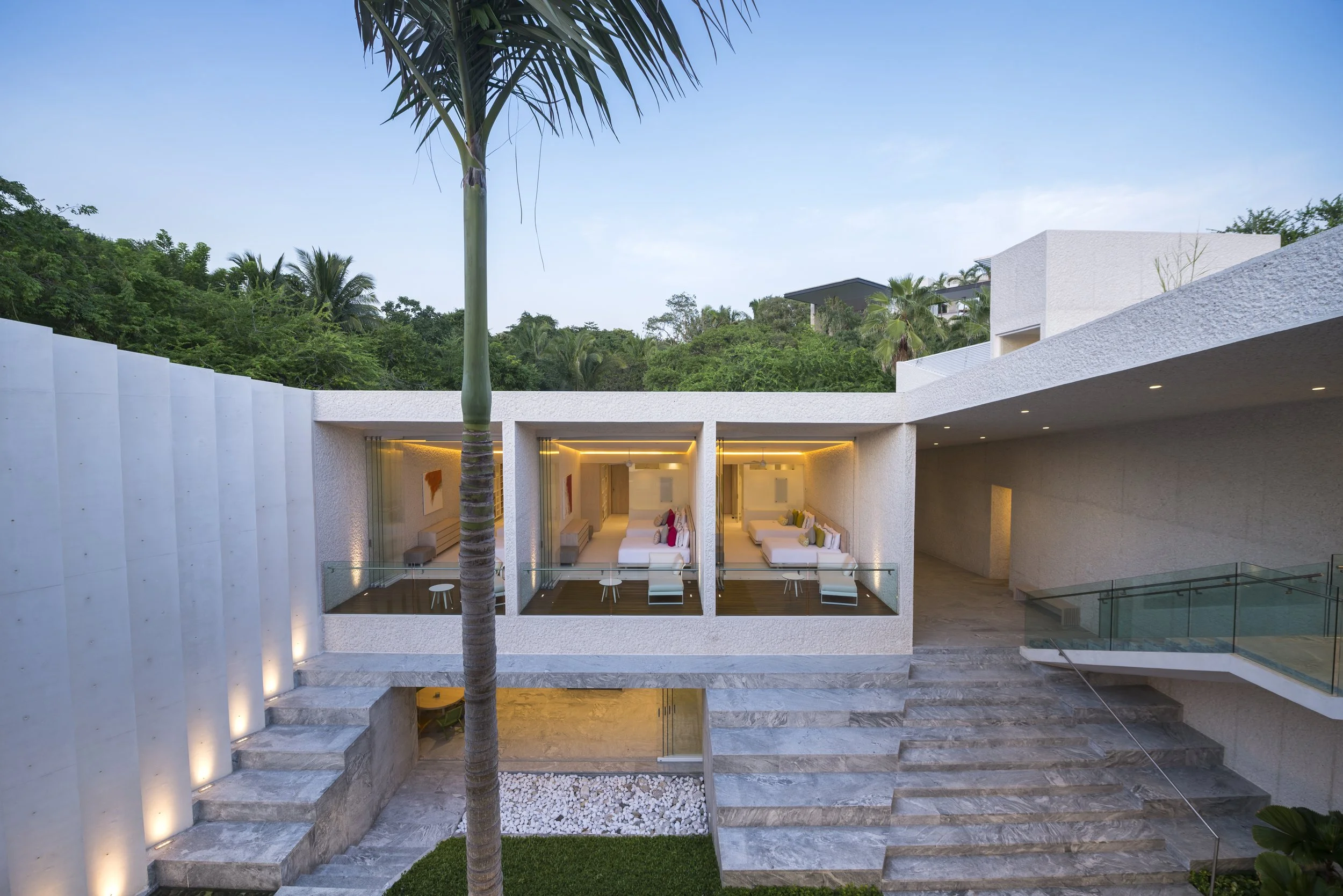 Modern residential building with open-air lounge area, large windows, white textured walls, and an outdoor staircase, surrounded by green trees and a blue sky.