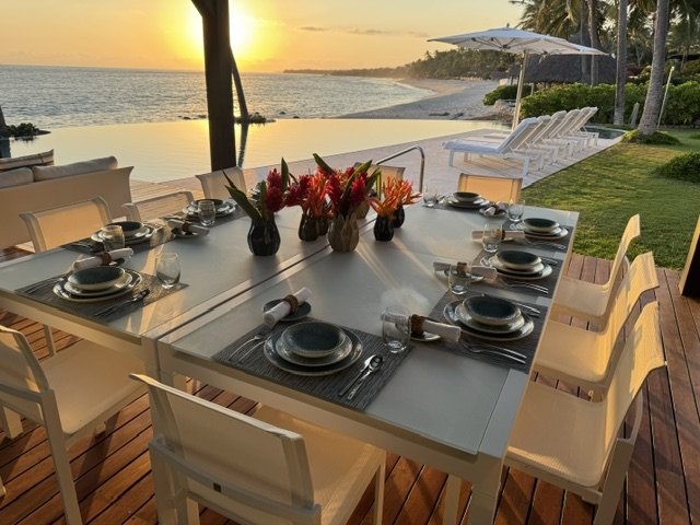 Dining table set with plates, glasses, and cutlery, decorated with colorful flowers, overlooking a beach at sunset with lounge chairs and umbrellas on the sand.