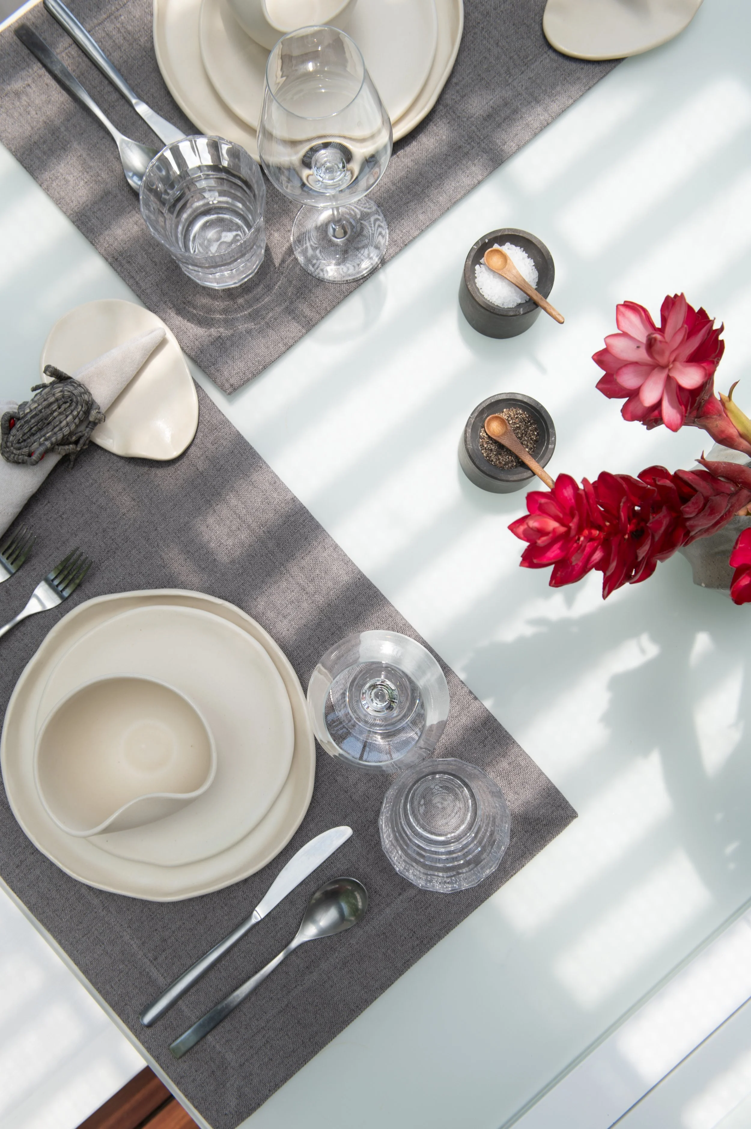 A table setting with white and beige dishes, crystal glasses, gray and white tablecloth, salt and pepper shakers with small wooden scoops, and pink flowers.