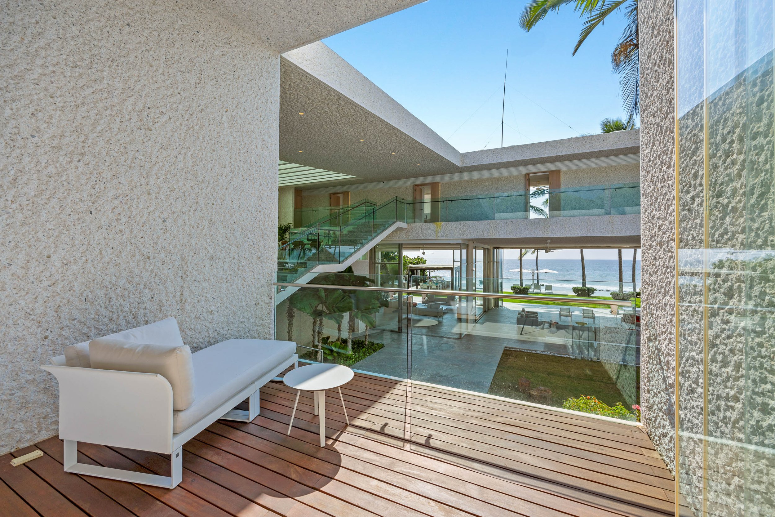 Modern interior space overlooking a beach with a deck area, white lounge chairs, and tropical plants, through a large glass window.