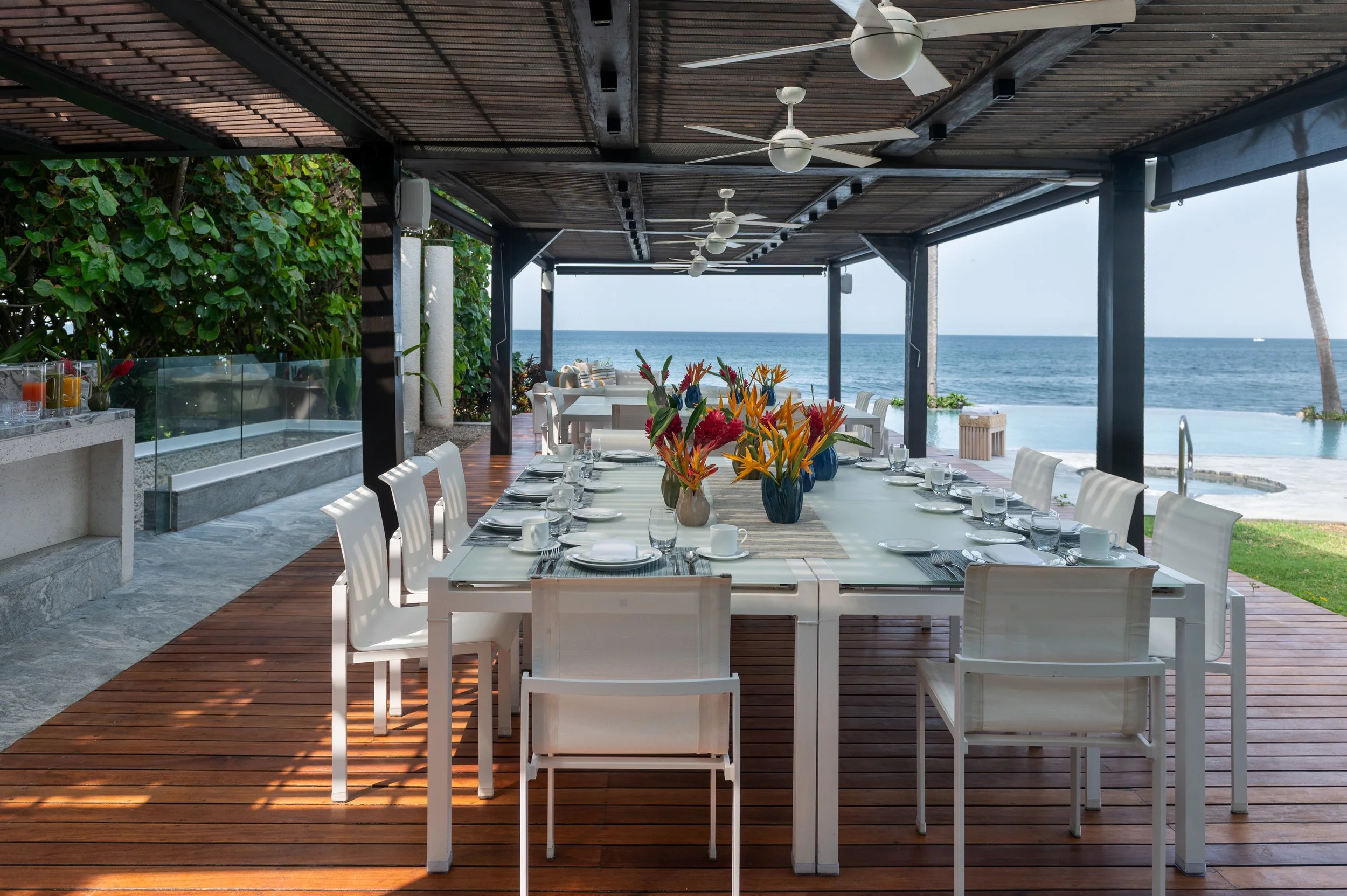 Outdoor dining area with a long table set with plates, glasses, and cutlery, decorated with colorful tropical flowers in vases, under a wooden roof with ceiling fans. The area overlooks the ocean with a clear blue sky and some greenery, including pal