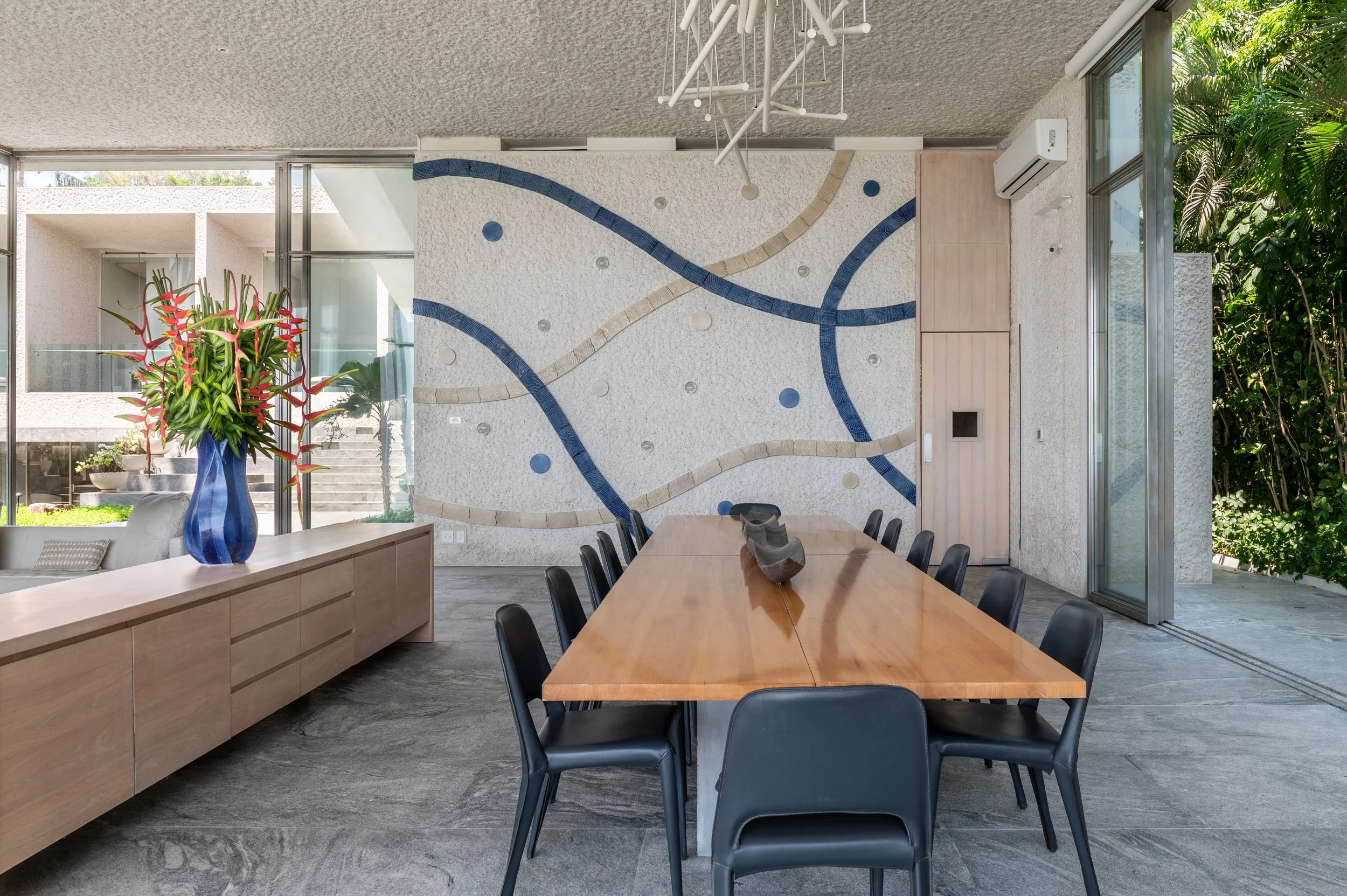 Modern dining room with large wooden table surrounded by black chairs, a wall with a textured surface and abstract blue and beige wall art, large windows with views of greenery, a blue vase with colorful flowers on a sideboard, and a contemporary lig