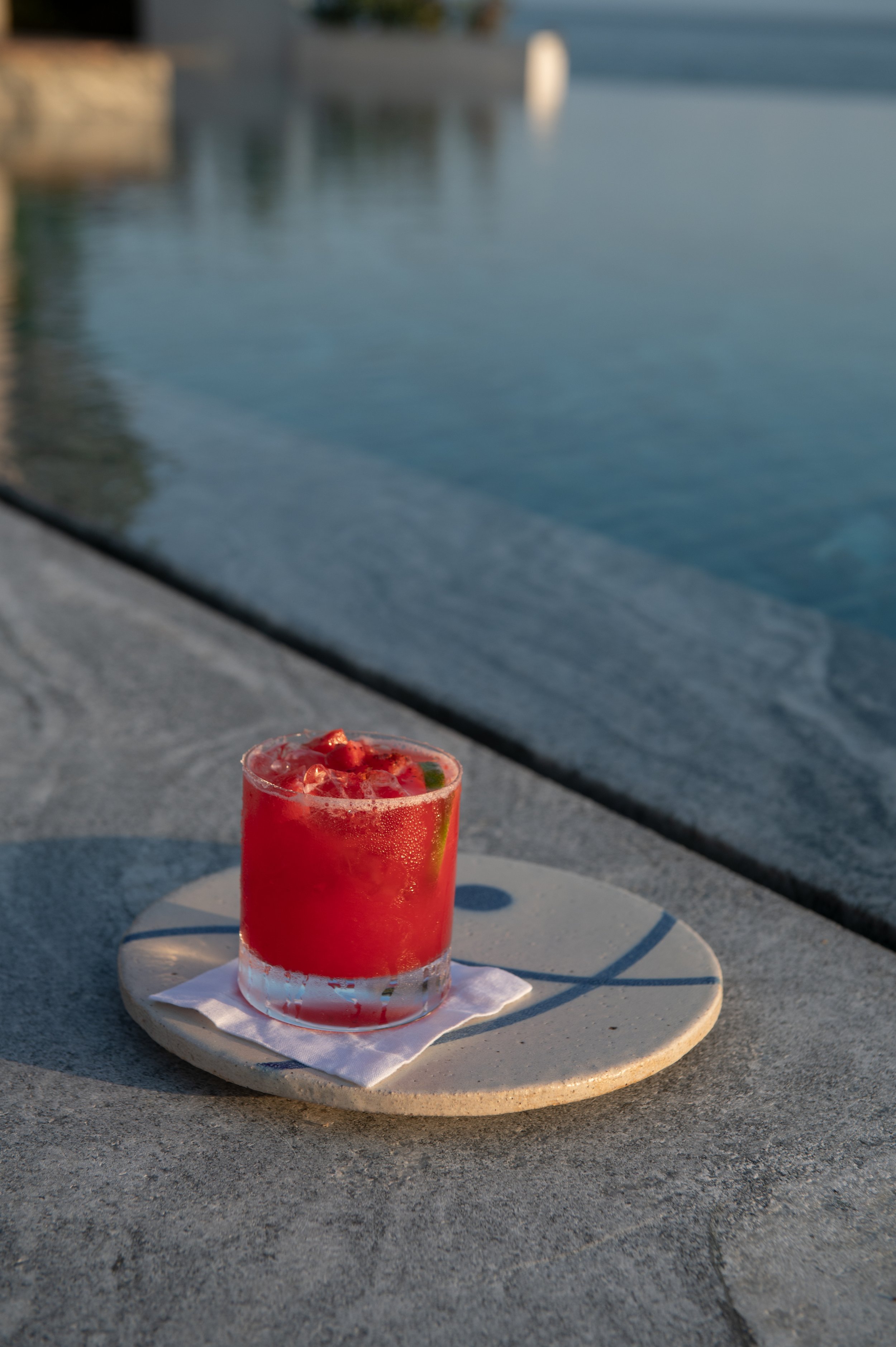 A glass of red cocktail with strawberries and lime on a white plate, set outdoors near a body of water during the day.