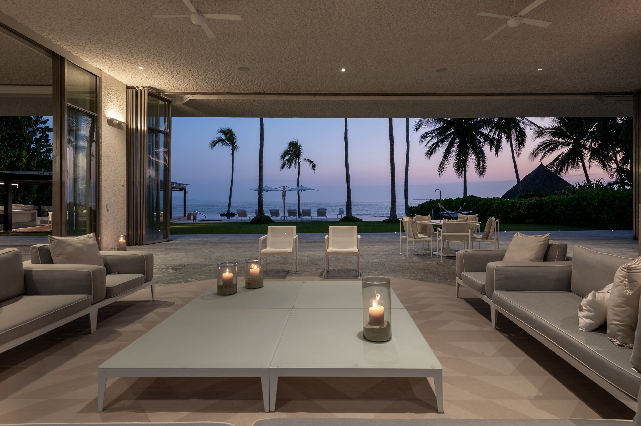 Indoor living area with white sofas, candles on white coffee tables, and view of palm trees and the ocean through open sliding doors at sunset.