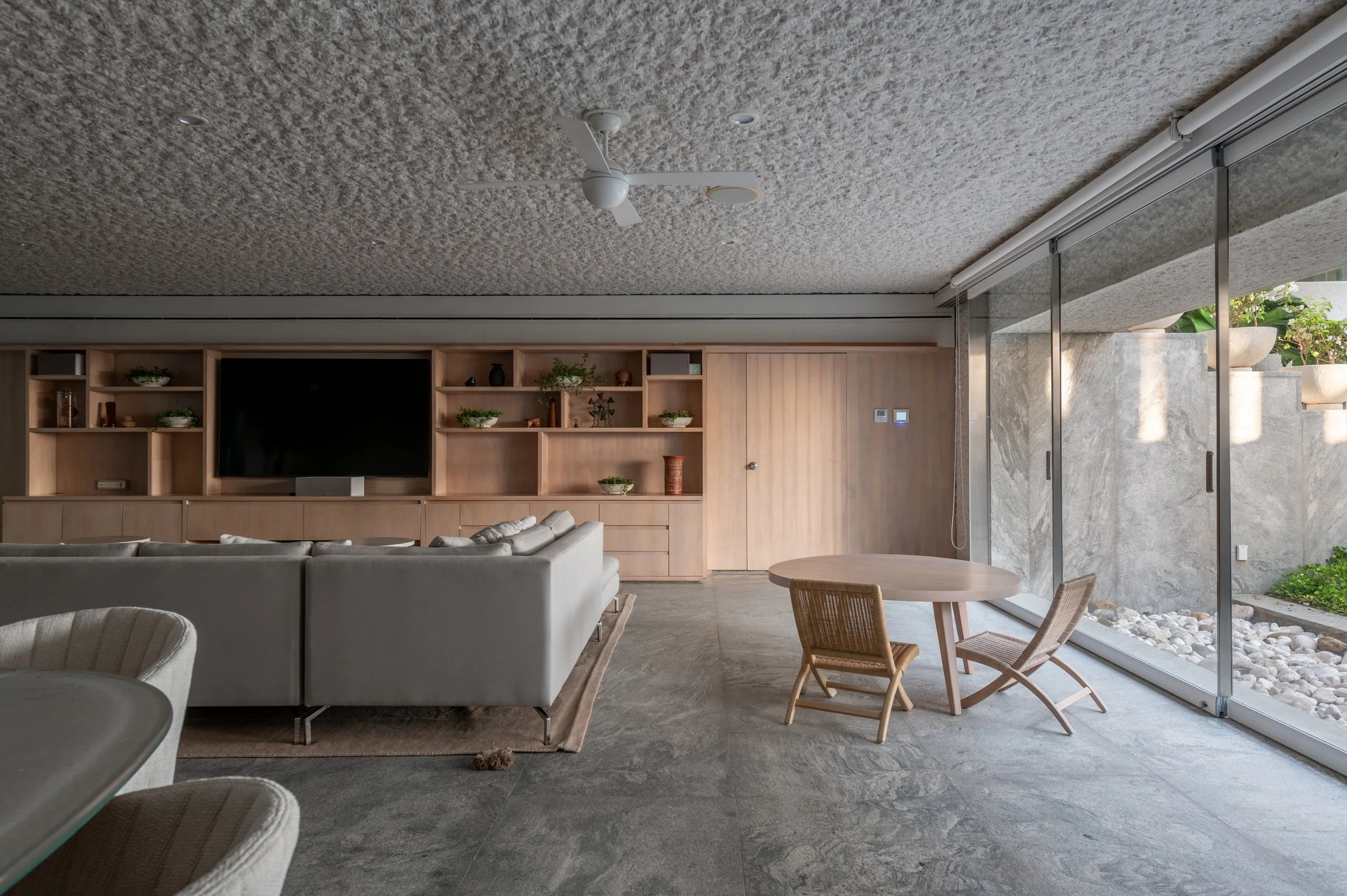 Living room with white sofas, a wooden bookshelf with decorative objects and plants, a round coffee table, and large glass sliding doors leading outside to a garden with rocks and plants.