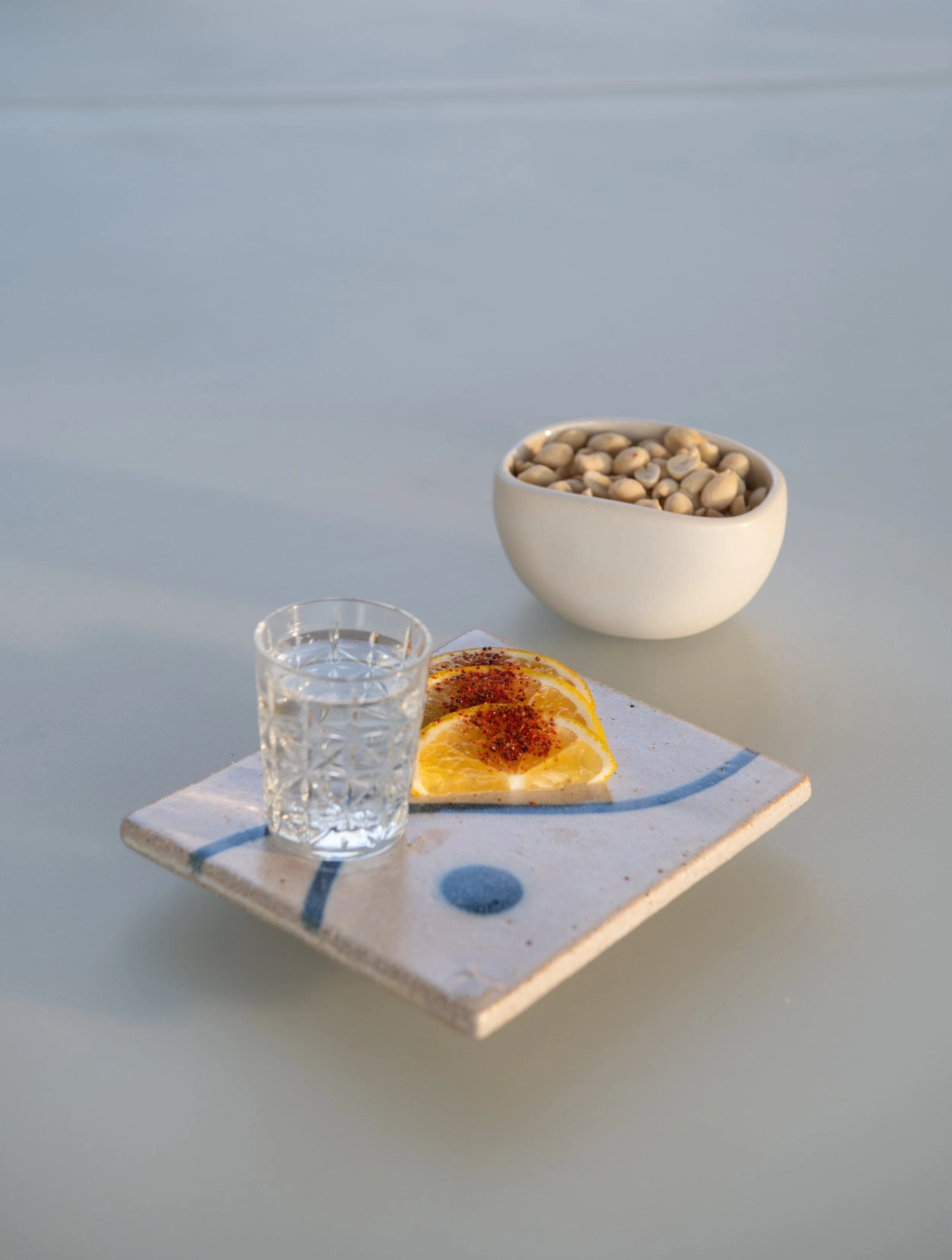 A white ceramic bowl filled with pine nuts on a white tiled serving tray, with lemon slices topped with chili powder and a glass of water in the foreground. In the background, there is another small glass container and a white textured surface.