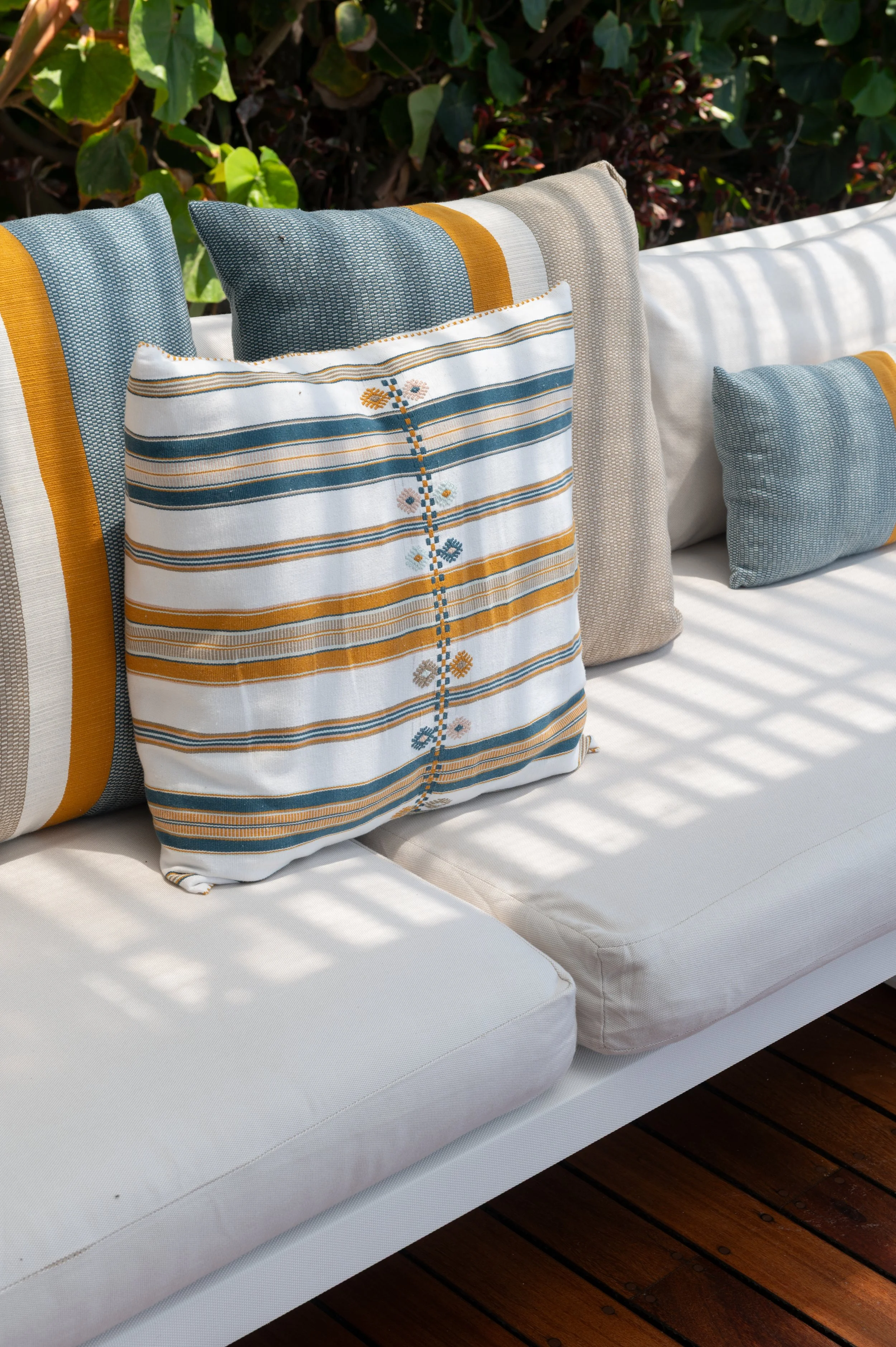 Outdoor seating area with white cushions and decorative pillows on a wooden deck, surrounded by green plants.