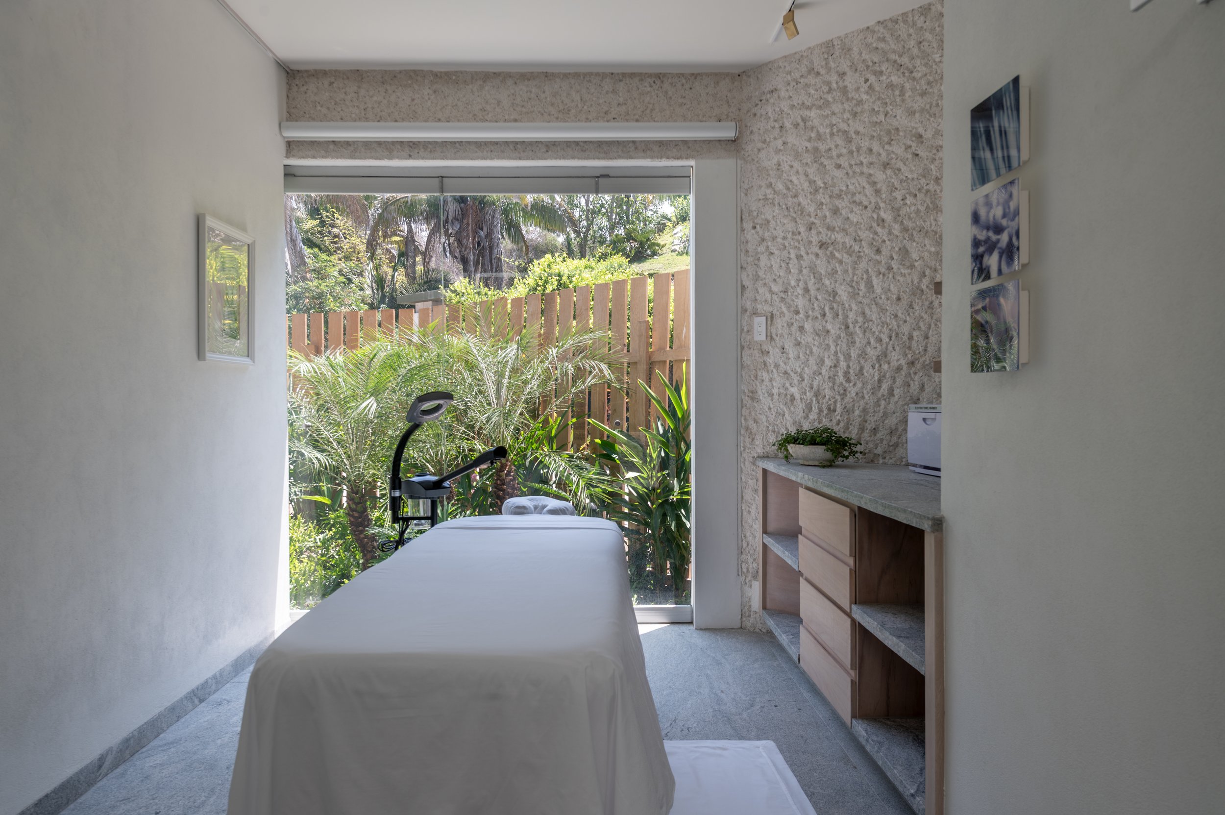 A bright room with a large sliding glass door leading to a lush outdoor garden with palm trees and a wooden fence. Inside, there is a massage bed with a white cover, a small black lamp, a wooden cabinet with open shelves, a plant, and a picture hangi