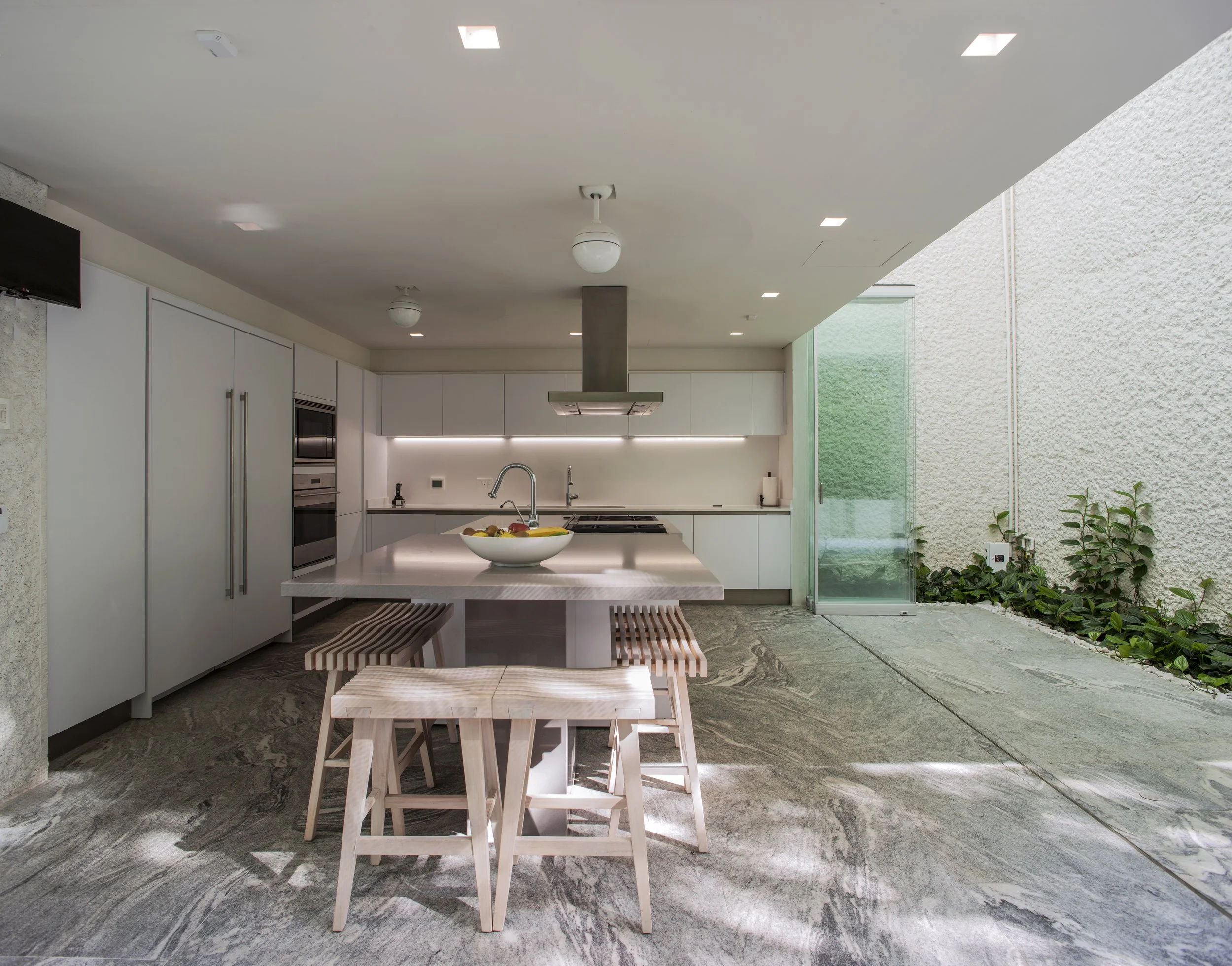 Modern kitchen with white cabinets, a white kitchen island, a fruit bowl, wooden stools, and an outdoor patio with plants.