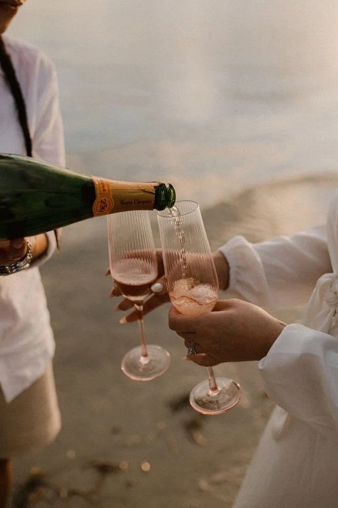 Two women clinking glasses of rosé champagne on a beach at sunset, with one woman pouring champagne from a green bottle.