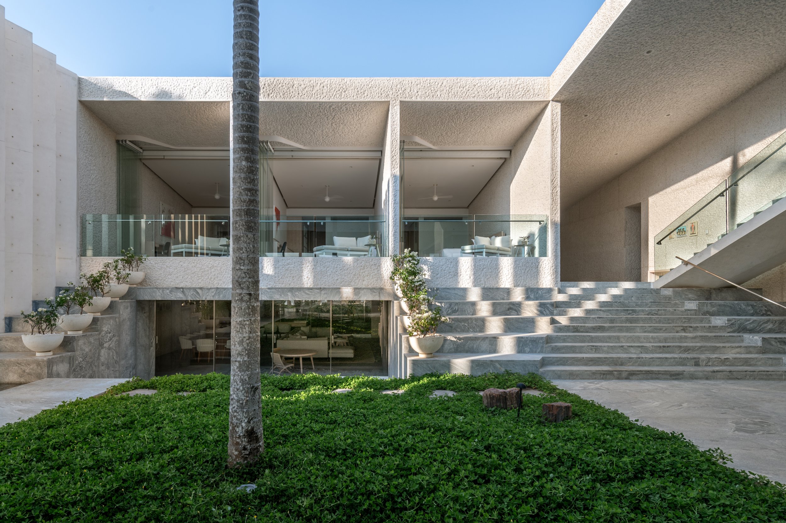 Modern multi-level building with large glass windows, concrete exterior, and exterior staircase, surrounded by green grass and potted plants with a tree in the foreground.