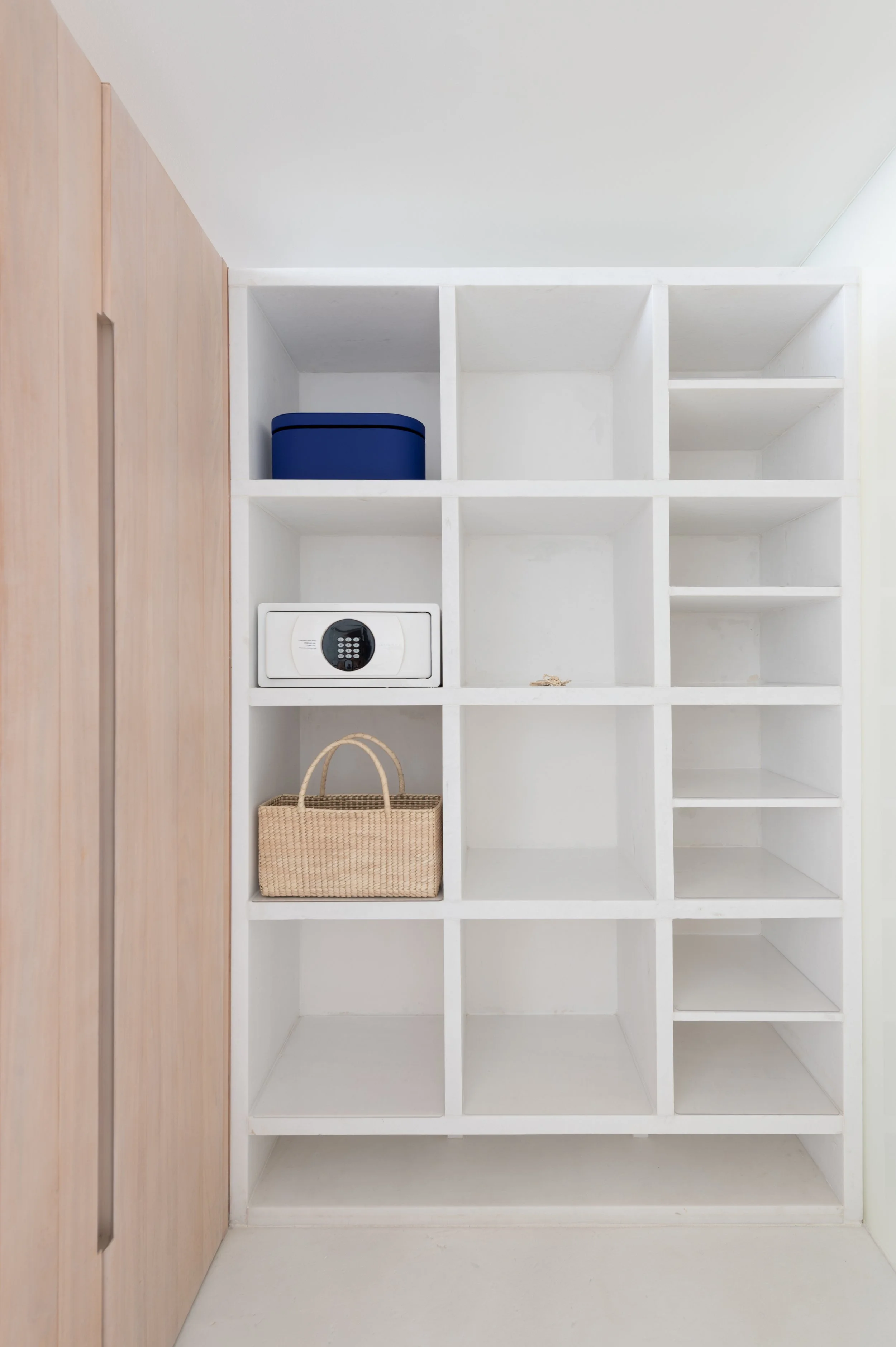 White shelving unit with empty compartments, a blue box in one compartment, a woven basket on the bottom shelf, and a wooden cabinet to the left.