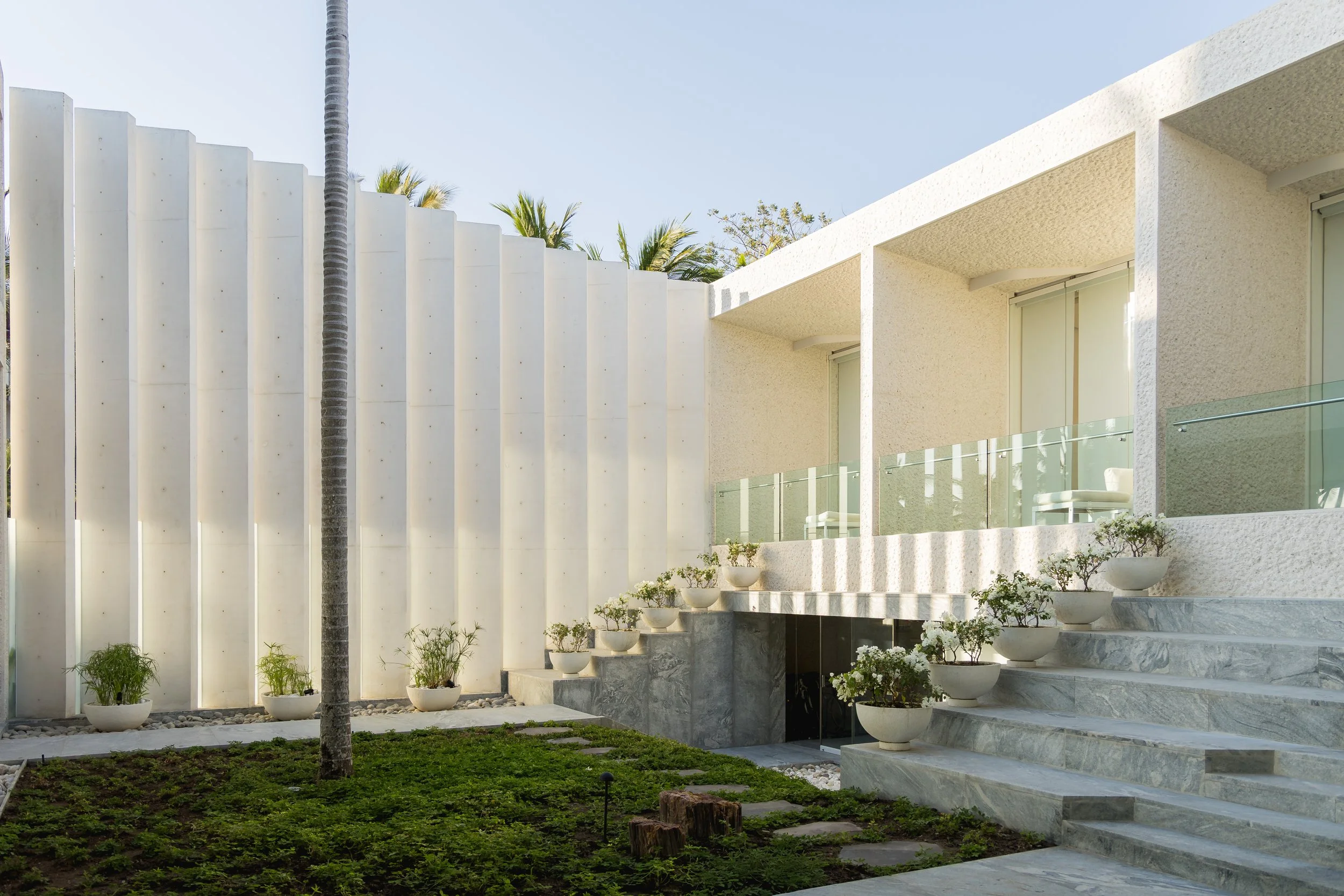 Modern residential building with white textured walls, large windows, and outdoor balcony with glass railing. Potted plants line the steps leading to the entrance, with a landscaped garden and a large palm tree nearby.