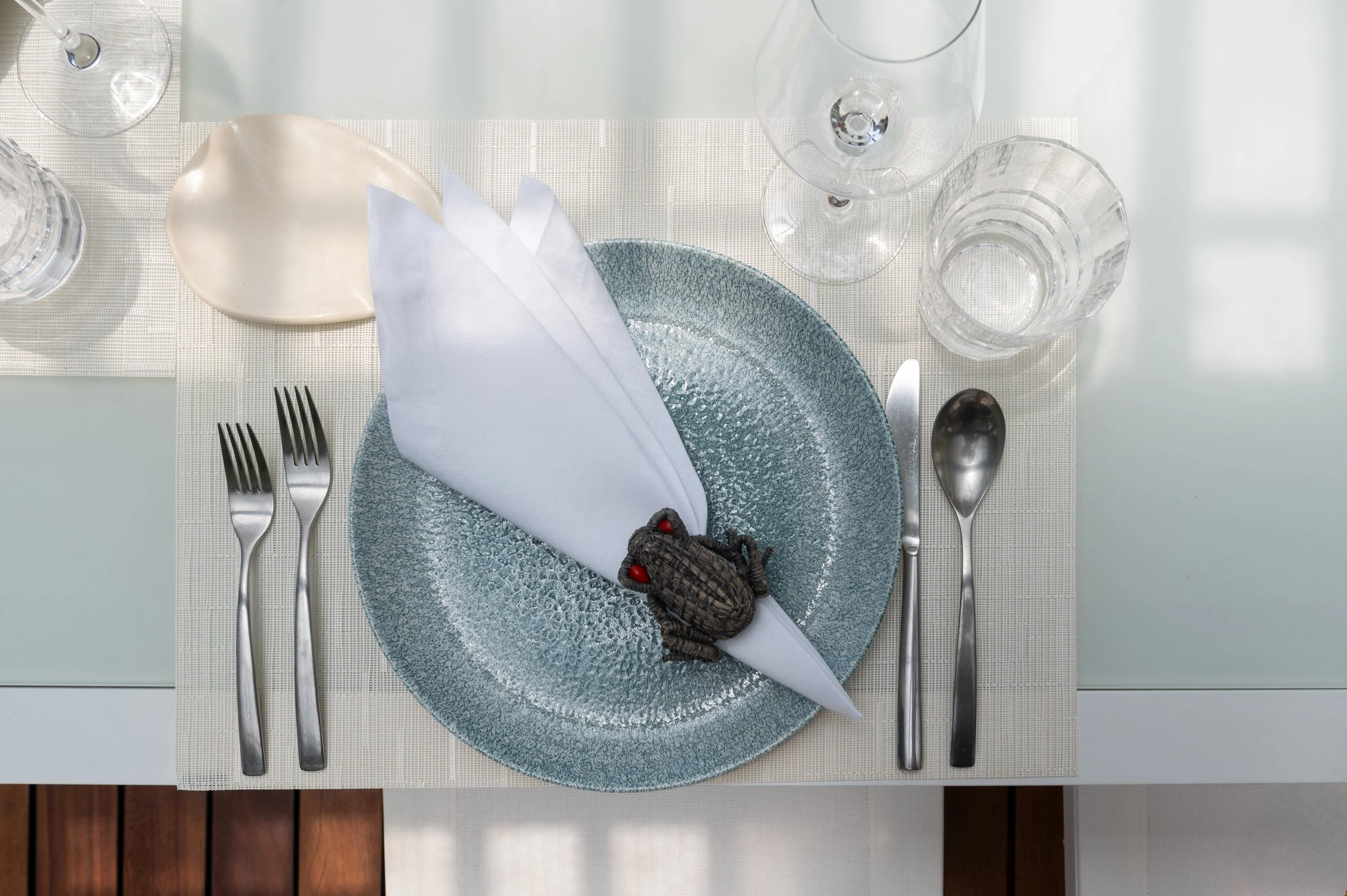 Restaurant table setting with a textured blue plate, a white napkin, silverware, and multiple glasses, including a wine glass and a tumbler. There is a decorative butterfly-shaped object on the plate.