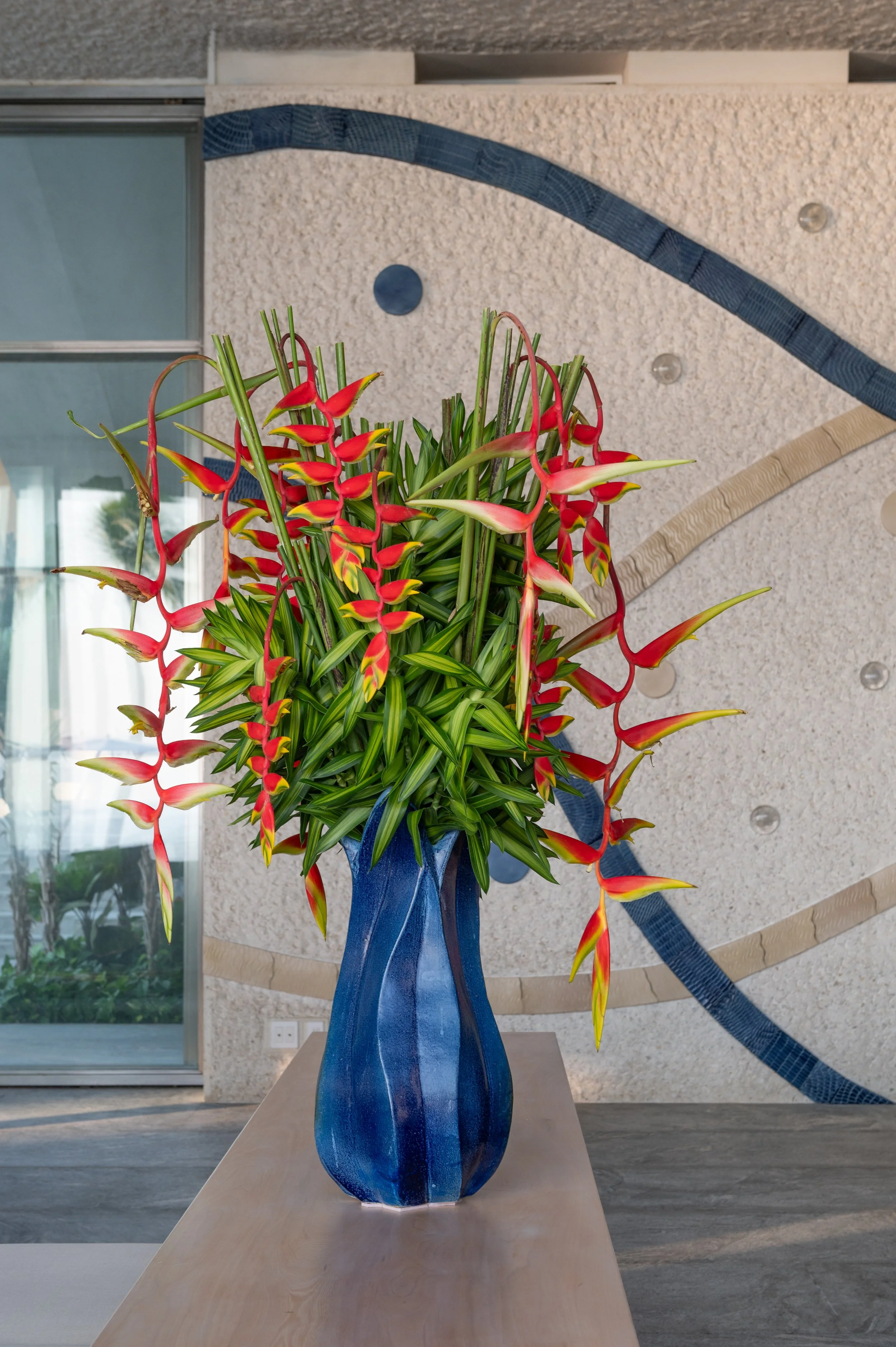 A blue ceramic vase holding a tropical flower arrangement with red and yellow heliconias and green foliage, placed on a wooden table indoors.