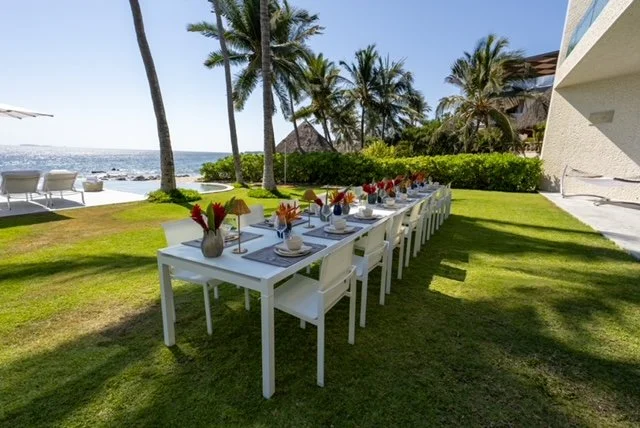 Long outdoor dining table set with white chairs, tableware, and colorful flower arrangements on a lush lawn near the beach with palm trees.