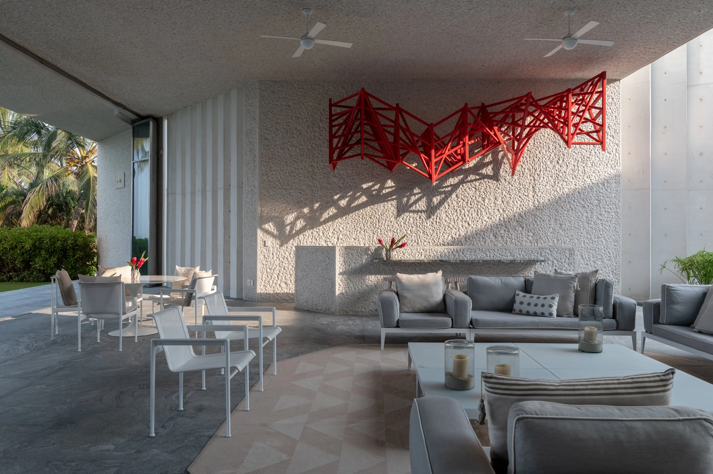 Modern outdoor living area with white chairs and sofas, a white table with candles, a textured white wall with a red geometric art installation, large windows, ceiling fans, and tropical plants outside.