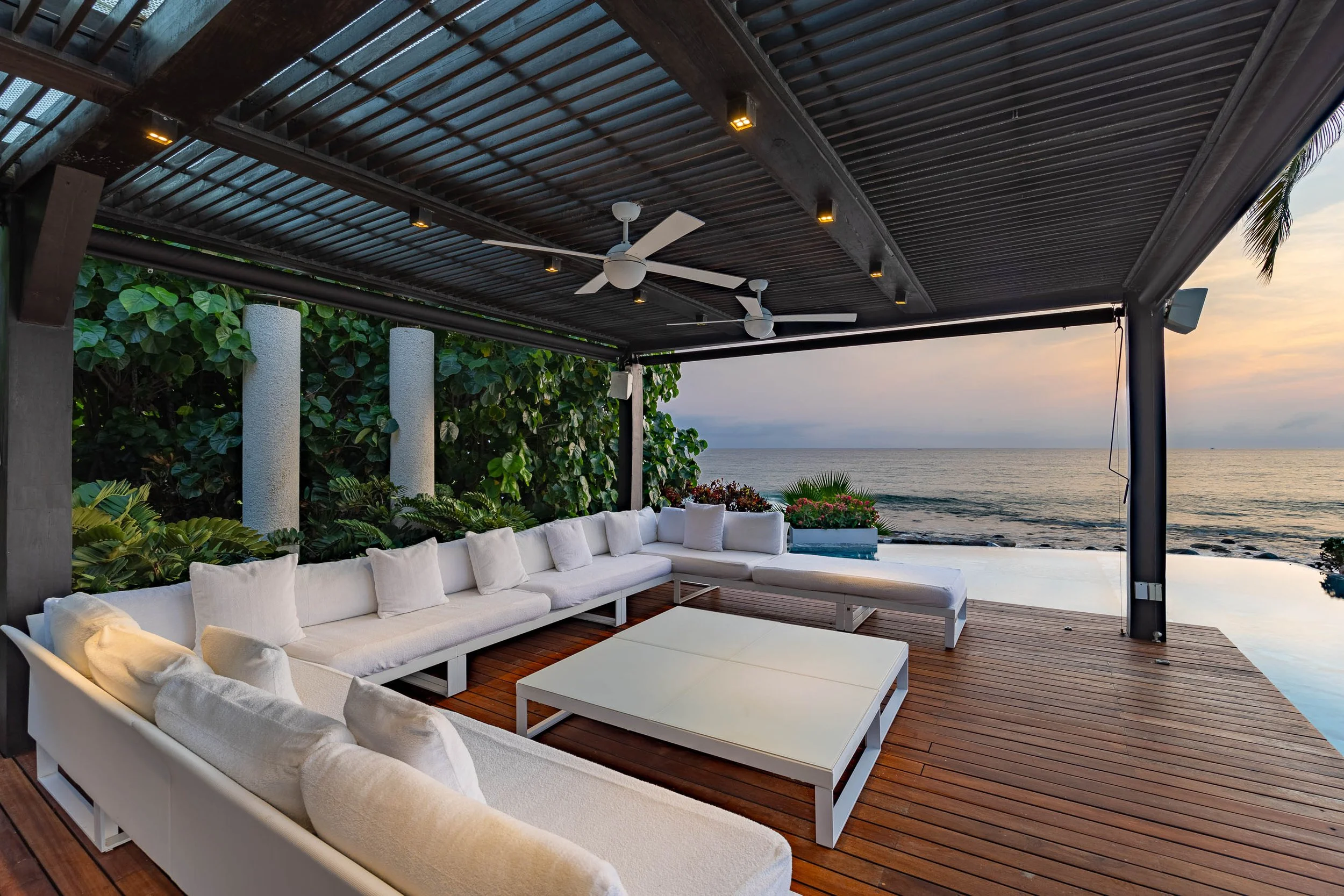 A modern outdoor patio overlooking the ocean with white cushioned seating, wooden flooring, a ceiling with rattan panels, and ceiling fans, surrounded by greenery and plants at sunset.