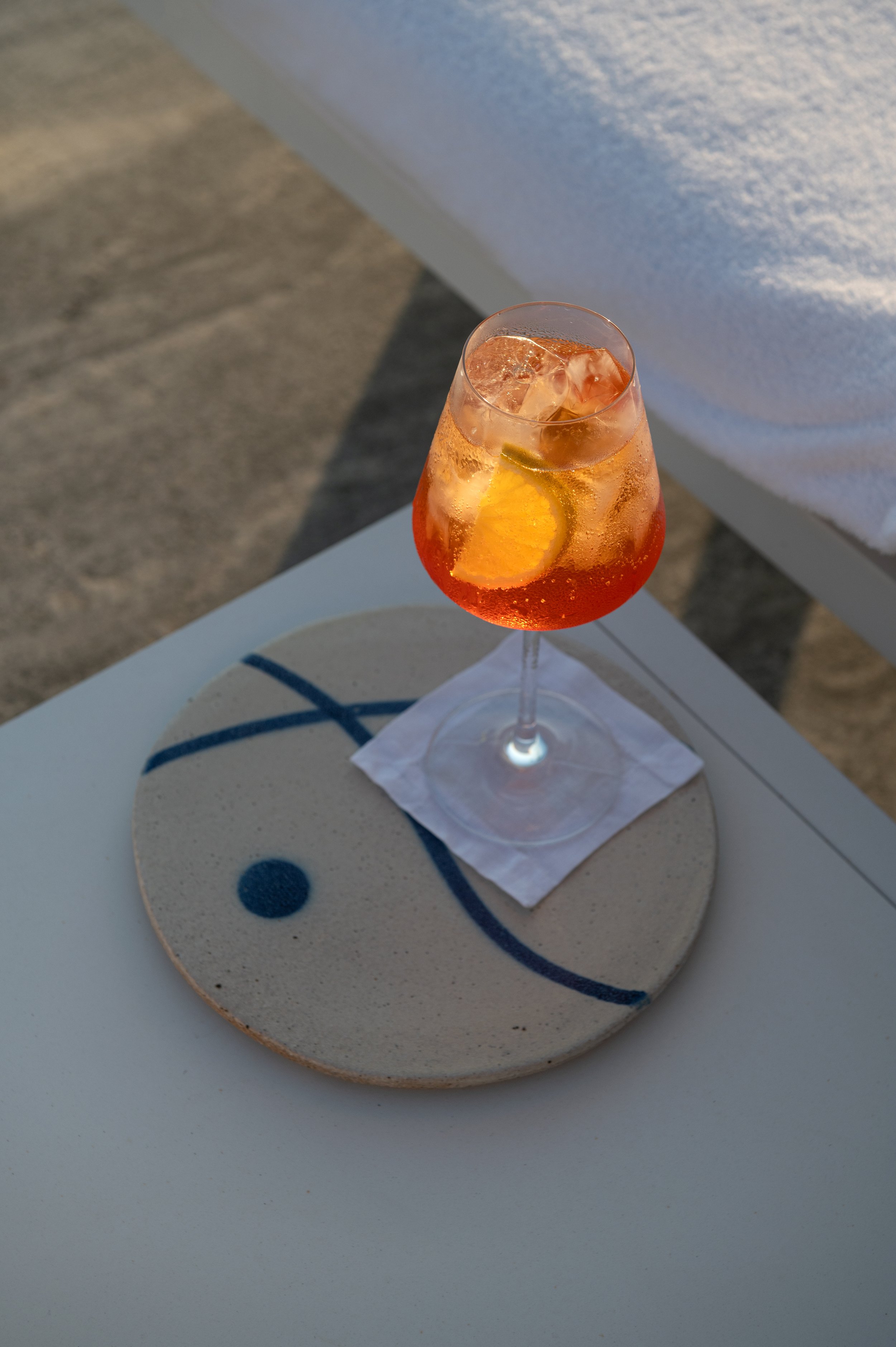 A tall glass of Aperol Spritz with ice, orange slice, and a slice of lemon, placed on a white table with a decorative ceramic coaster featuring blue geometric designs. The drink appears to be outdoors with sunlight and a textured ground in the backgr