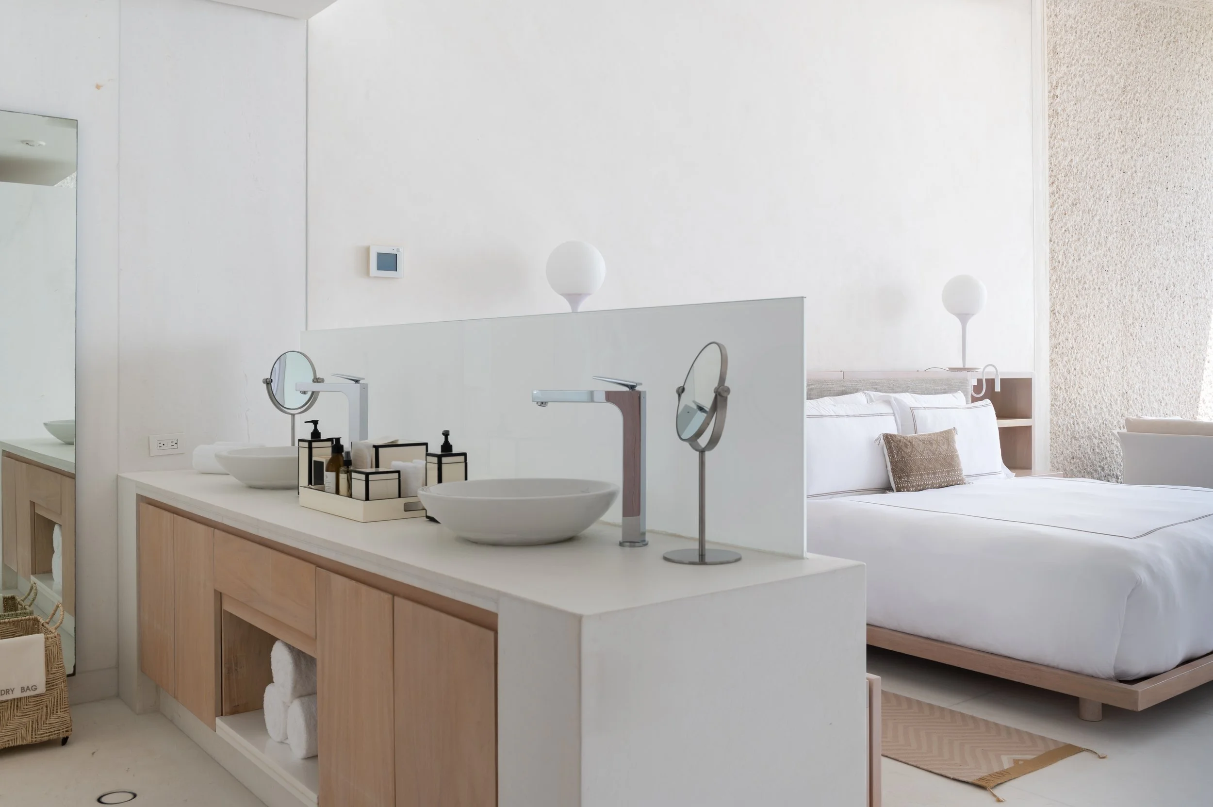 A modern bathroom with two white vessel sinks, two round mirrors, and black soap dispensers on a light wooden vanity. Behind the vanity is a bedroom area with a white bed, pillows, and two white bedside lamps. The room has white walls and a textured 