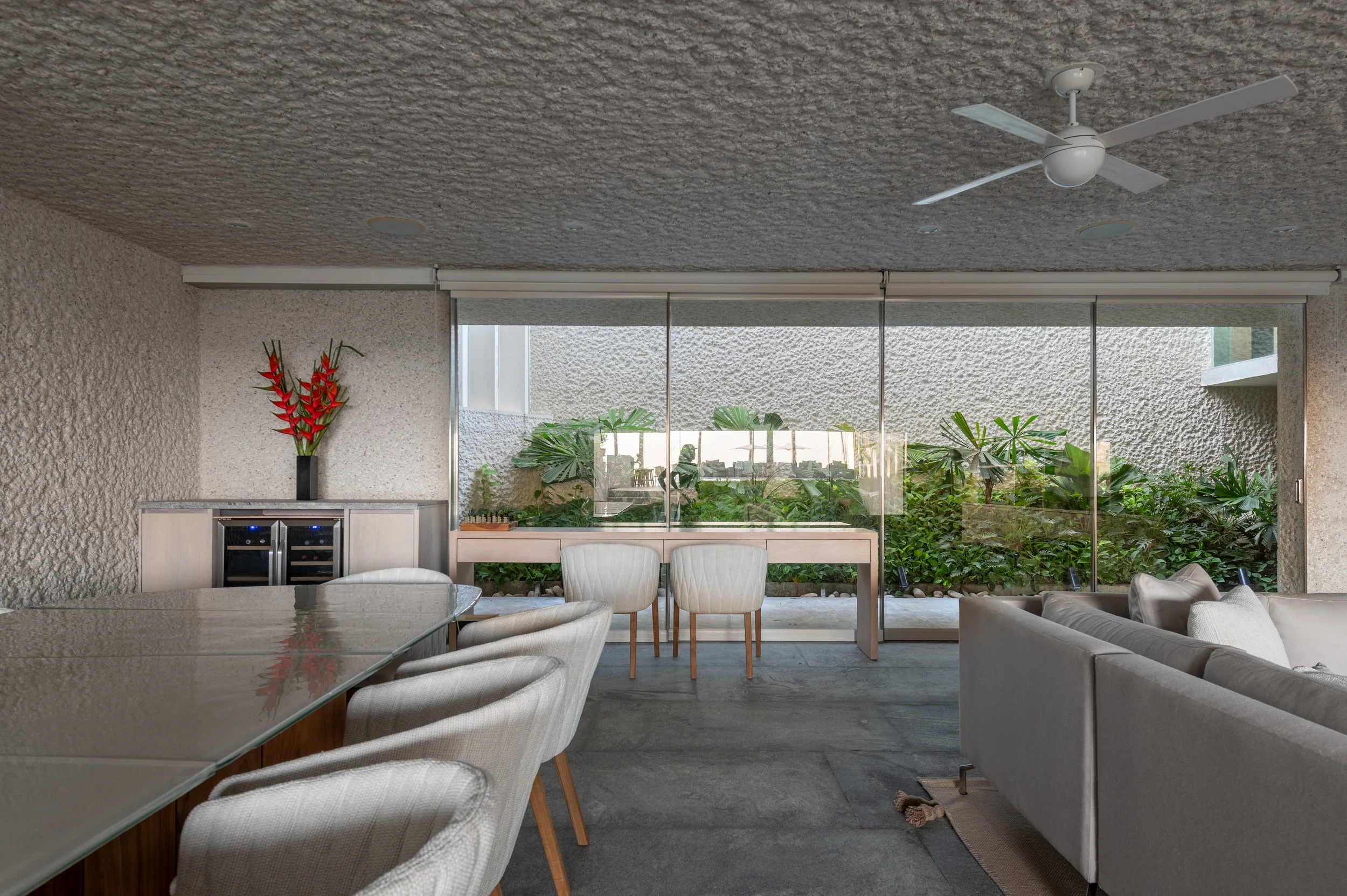 Modern living room with a textured ceiling, ceiling fan, large sliding glass doors, tropical plants outside, a dining area with cream-colored chairs, a glass-top table, and a white sofa.