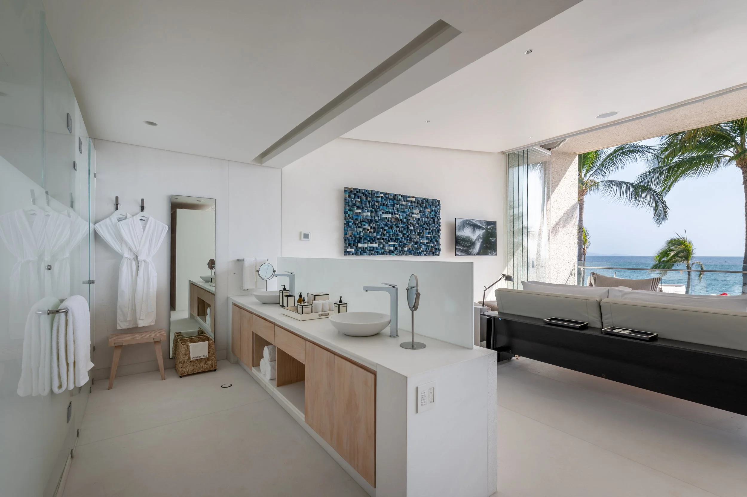 Modern bathroom with a view of the ocean featuring white walls, a wooden vanity with two vessel sinks, towels, a mirror, and an open balcony with palm trees and ocean in the background.