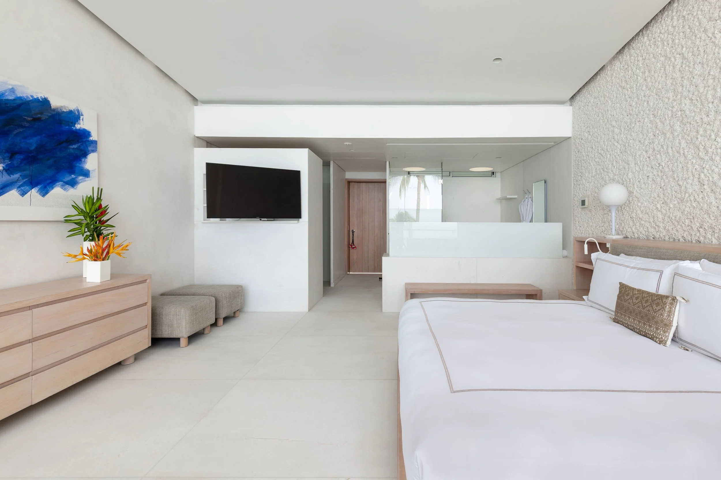 Modern living space with white walls, a bed with white linens, a wooden dresser with a potted plant and orange flowers, a flat-screen TV, and a textured wall in the background.