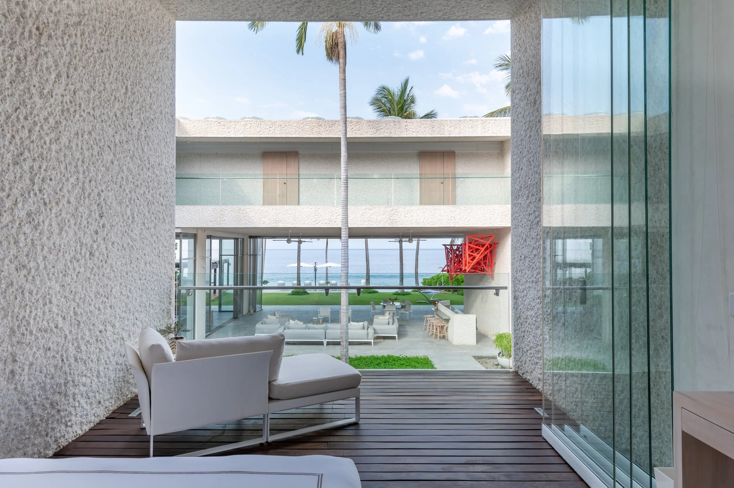 View from inside a modern building overlooking a patio with white outdoor furniture, palm trees, and the ocean in the background.