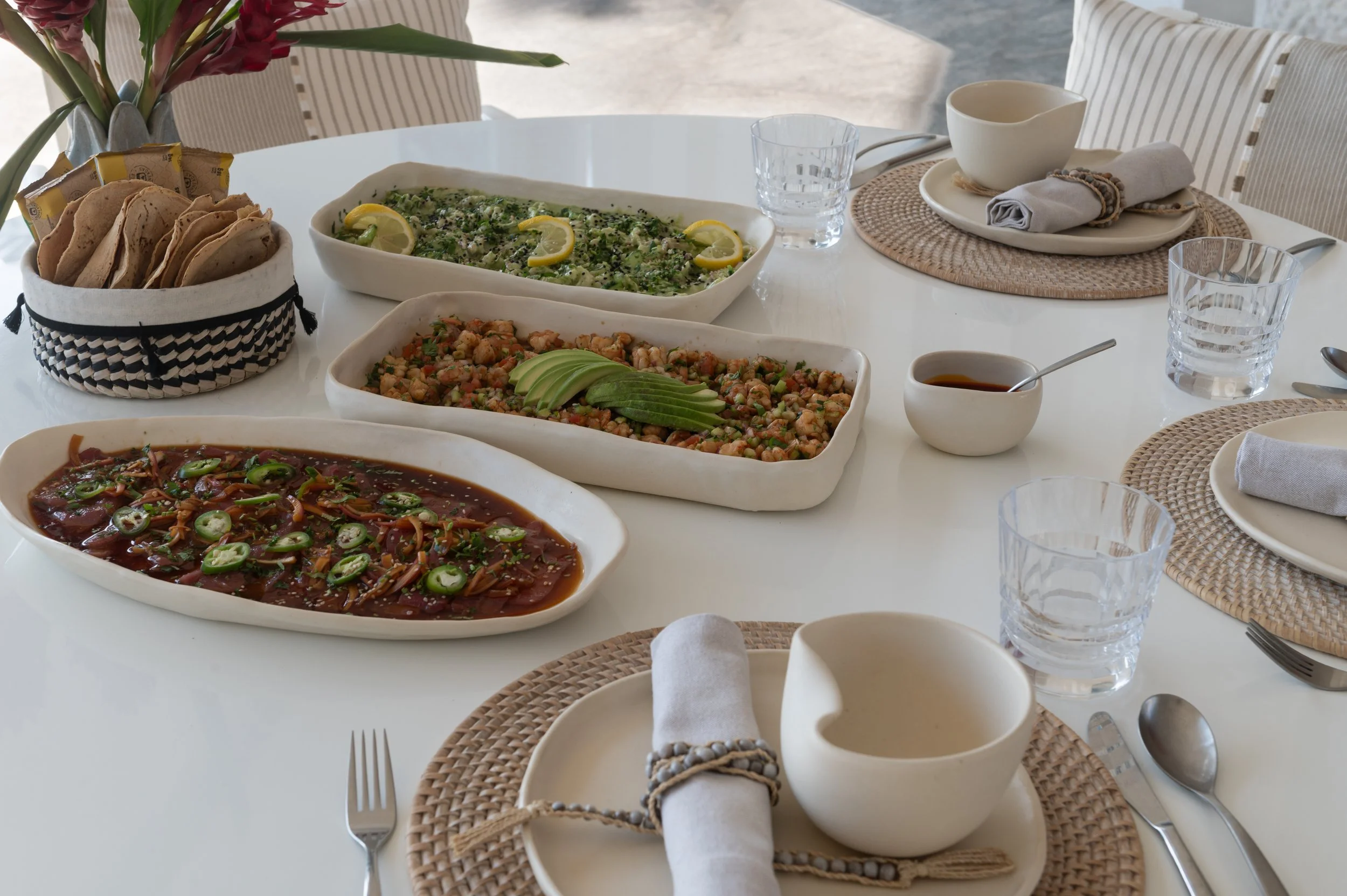 A white dining table set with various dishes including a plate of soup, a dish of cooked chickpeas with avocado slices, a bowl of pico de gallo, and a baked dish with lemon slices, surrounded by plates, glasses, and napkins, with a woven placemat and