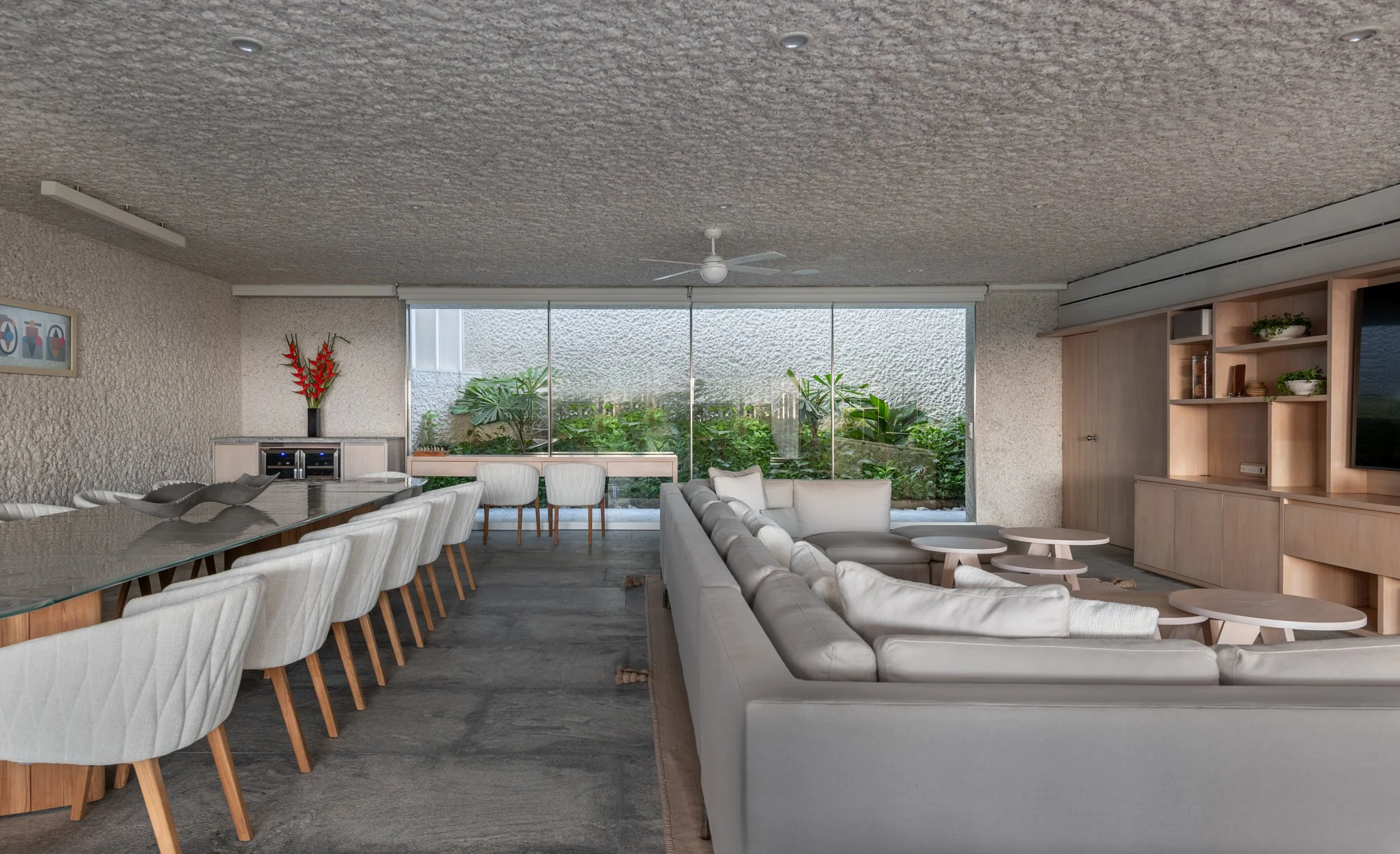 Living room with large window overlooking a garden, featuring white sofas, a glass dining table with chairs, and wooden shelving with decor items.