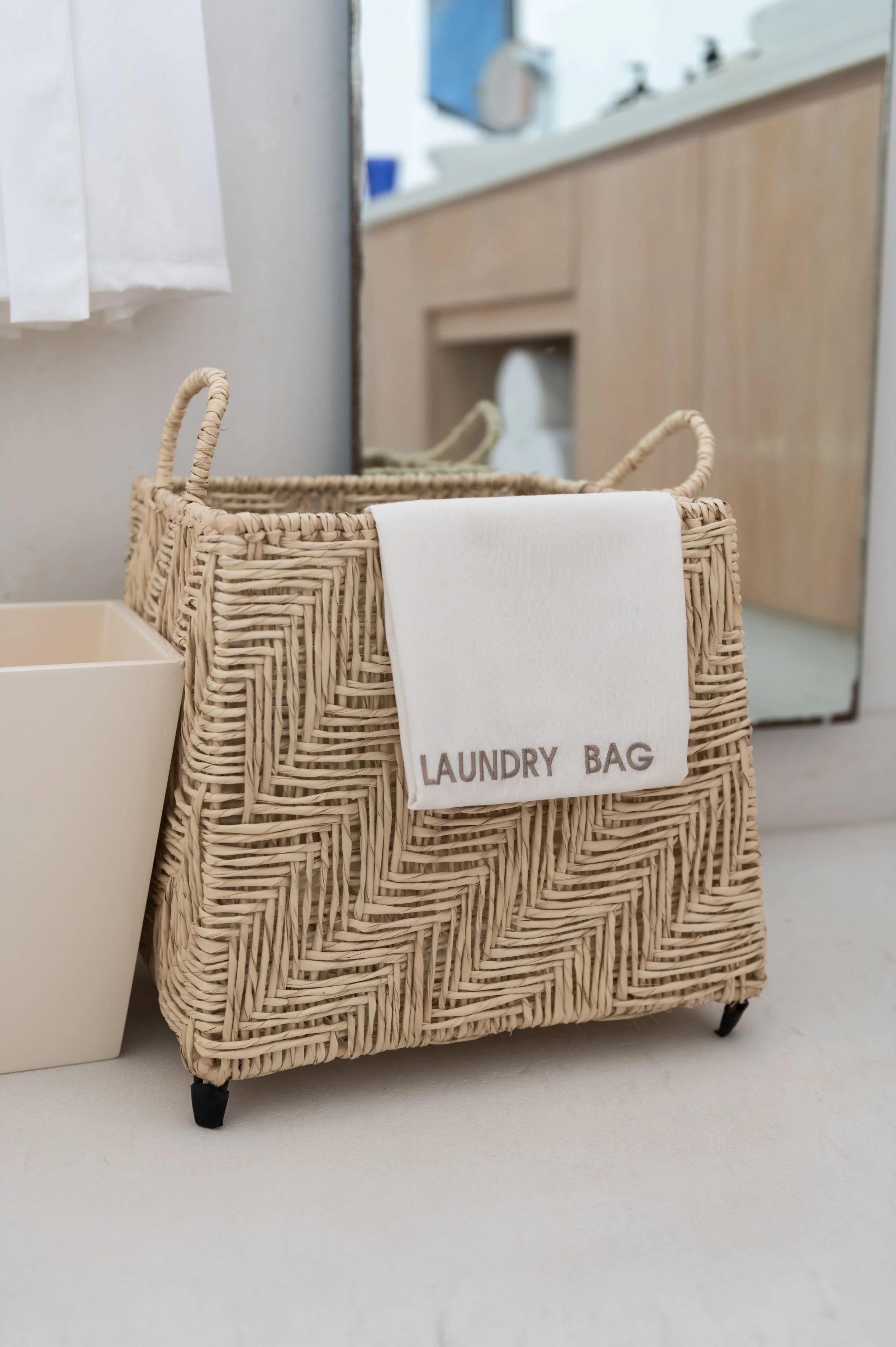 Wicker laundry basket with a cloth label reading "LAUNDRY BAG," on a light-colored surface with a portion of a bathroom visible in the background.