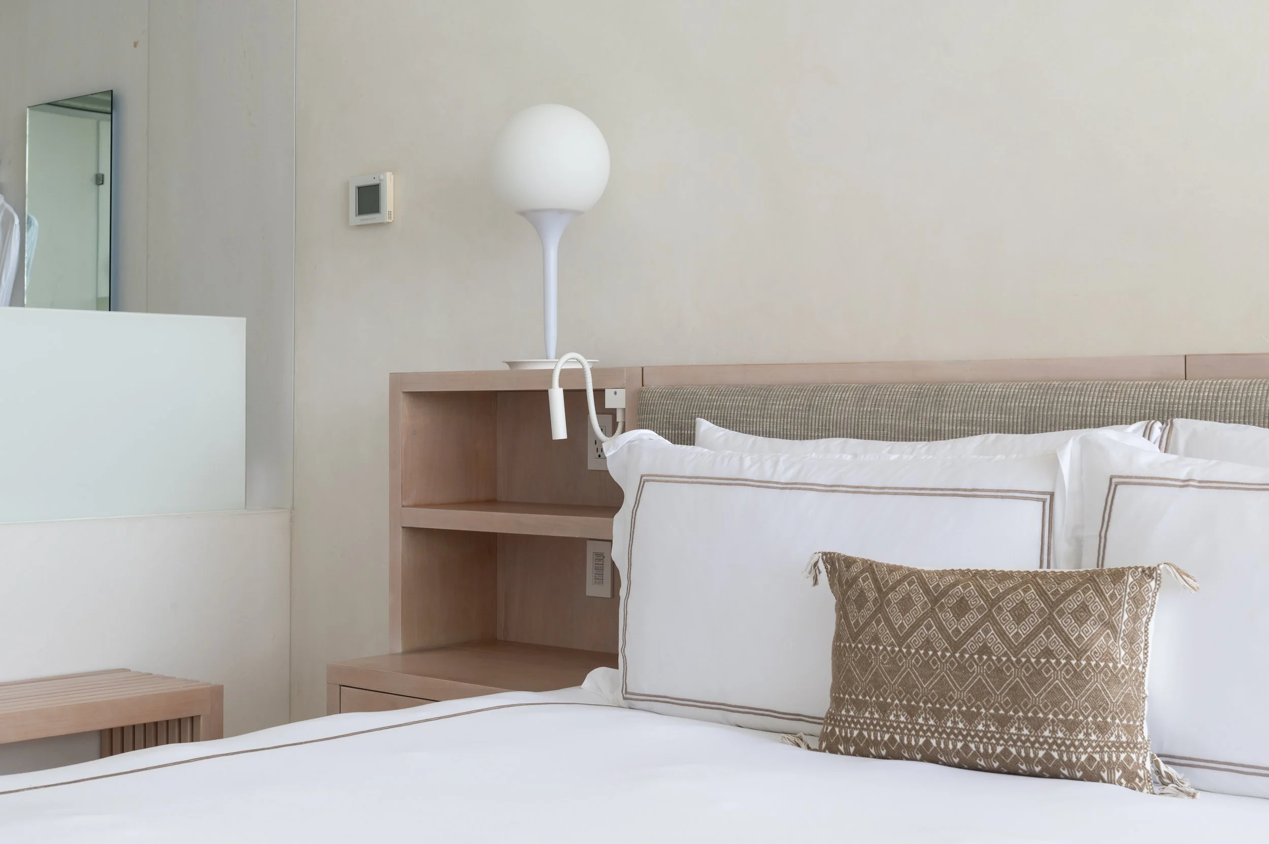 A hotel room bed with white bedding, a decorative pillow, a wooden headboard, and a side table with a white modern lamp attached to the wall. There's a mirror and a thermostat on the wall.
