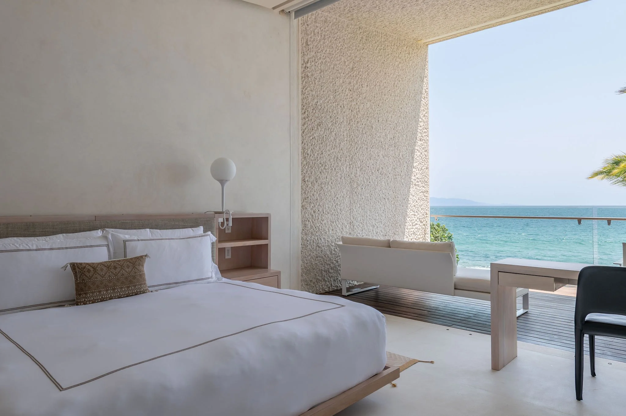 A bedroom with a large bed, decorative pillow, wall-mounted light, and ocean view through an open balcony with a white chair and table, palm trees, and a mountain in the distance.