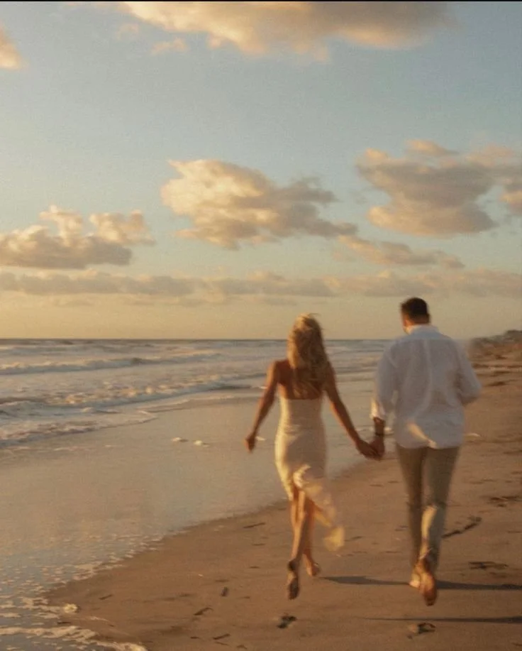 A couple holding hands and walking along the sandy beach at sunset, with clouds in the sky and ocean waves nearby.