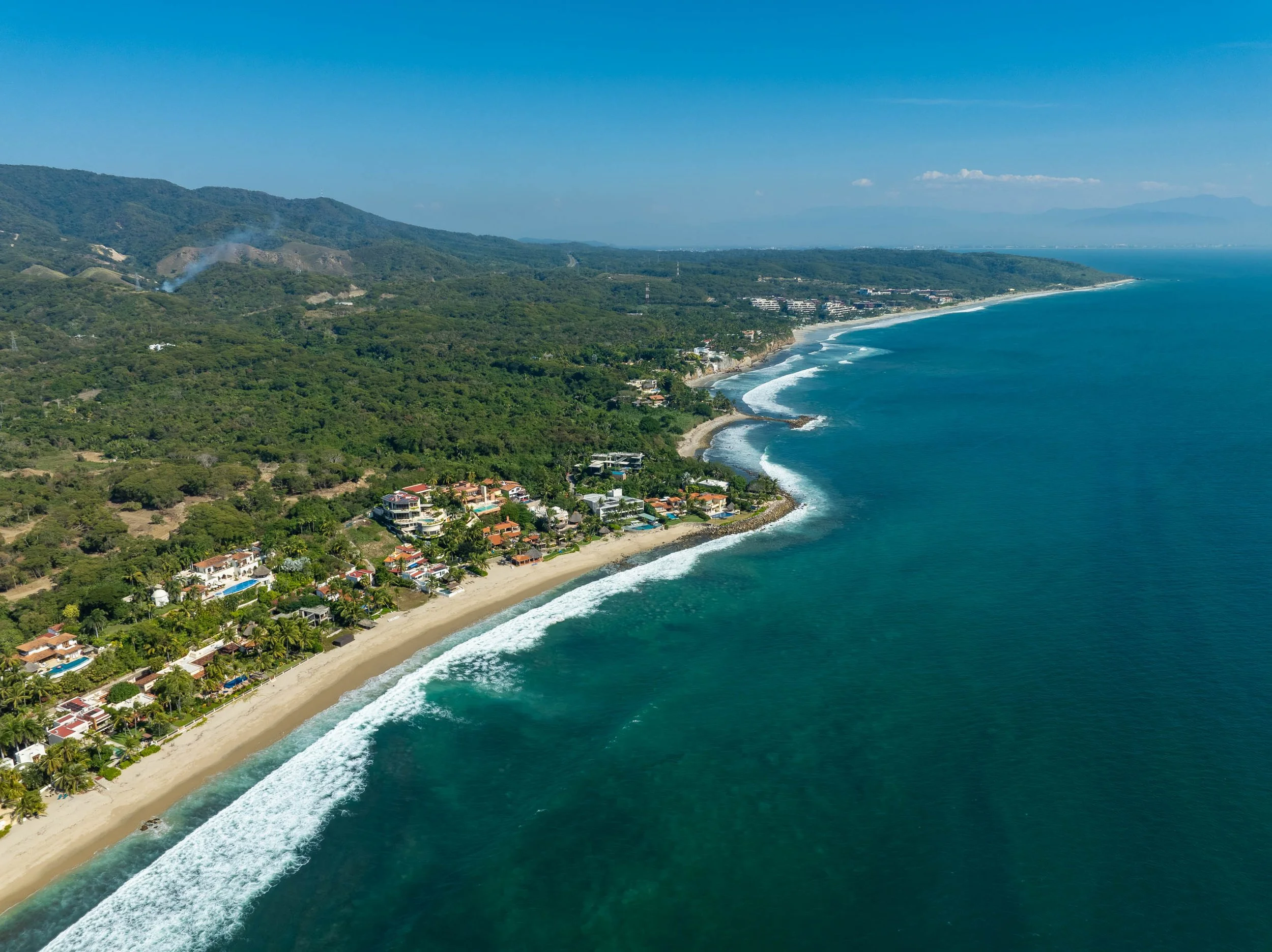 Aerial view of a coastal landscape with clear blue ocean waves hitting a sandy beach, green hills, and residential buildings along the shoreline under a clear blue sky.