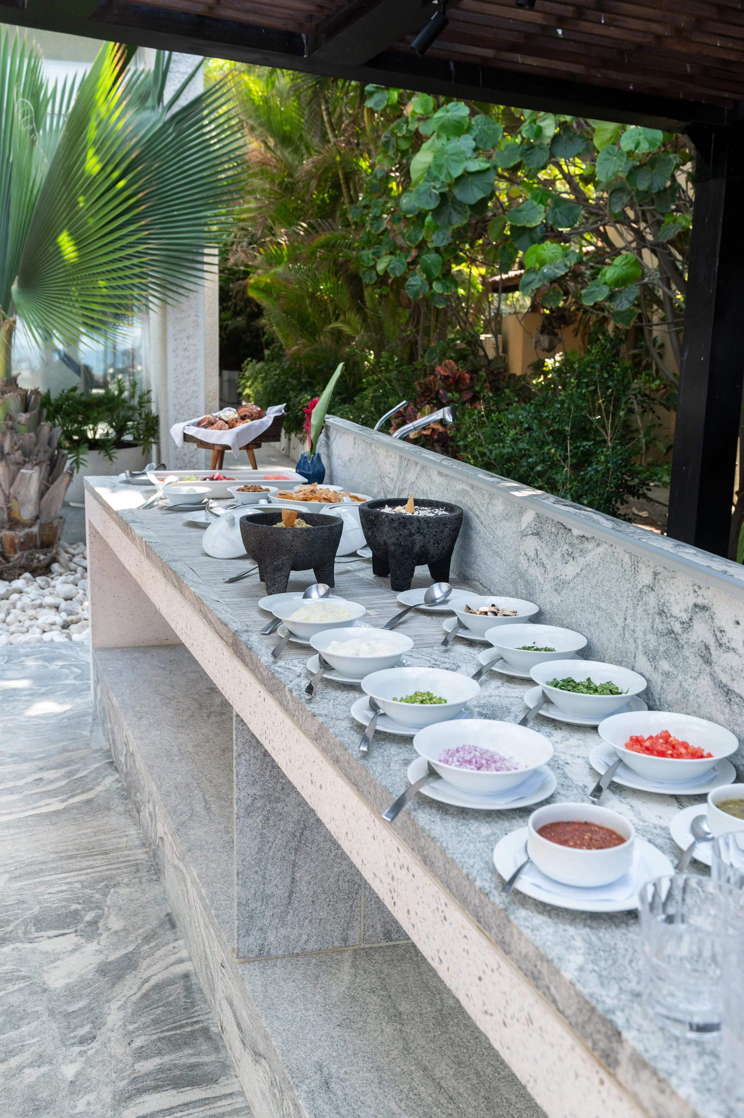 Outdoor buffet table with various bowls of toppings and ingredients, set against lush green plants and trees with a modern house in the background.