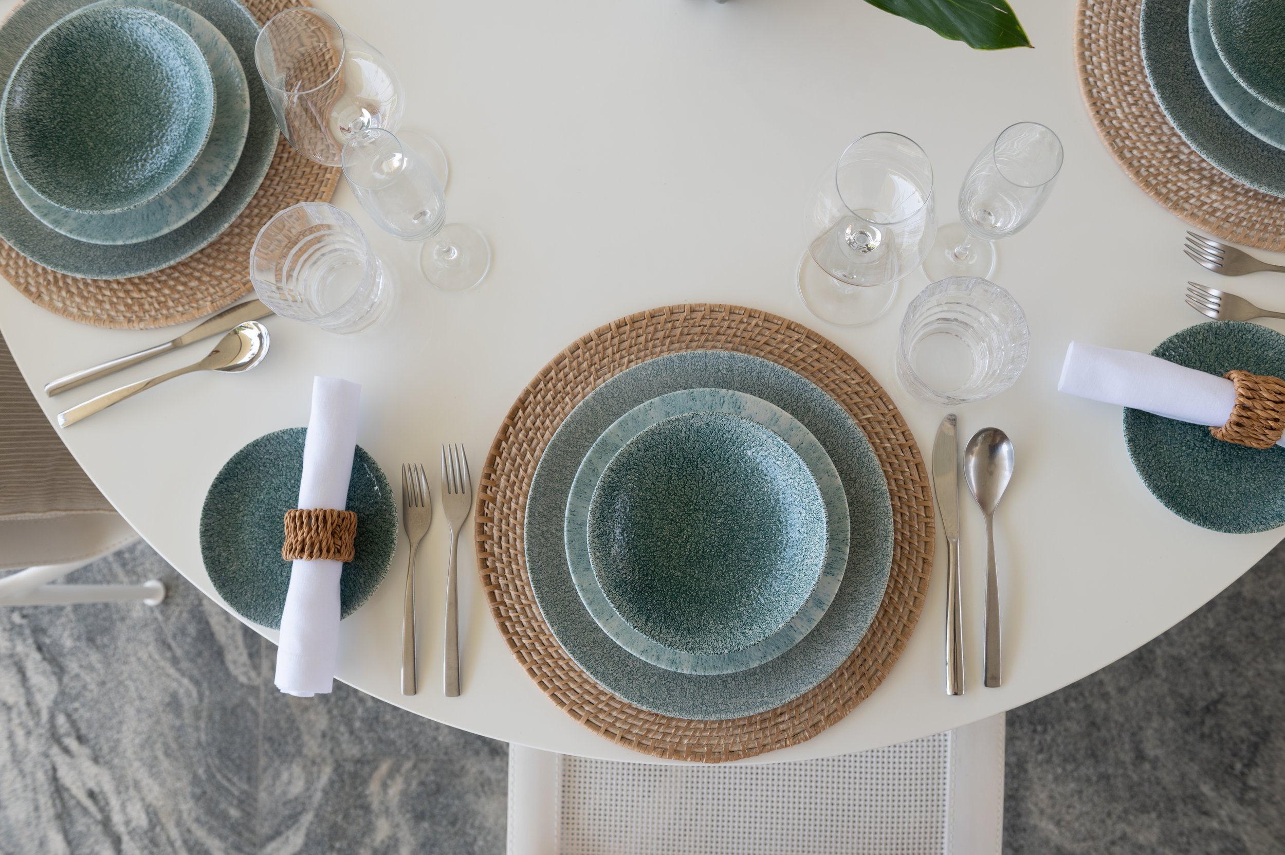 Table setting with teal plates, clear glassware, silver utensils, white napkins with woven napkin rings, rattan placemats, and a green plant in the background.