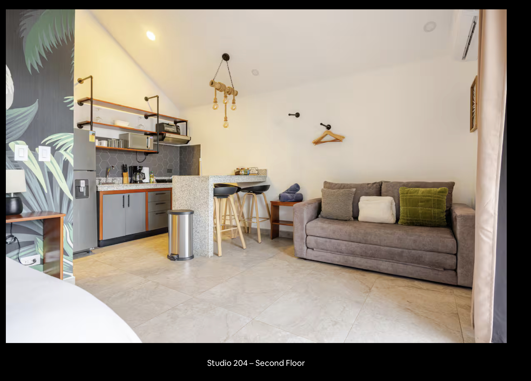 A compact studio apartment living area featuring a kitchen with gray cabinets and hexagonal black backsplash, a gray sofa with cushions, a small wooden side table, and a bar area with bar stools. The wall has a leafy mural design, with hooks and a hanger on the white wall. There is a hanging light fixture with exposed bulbs.