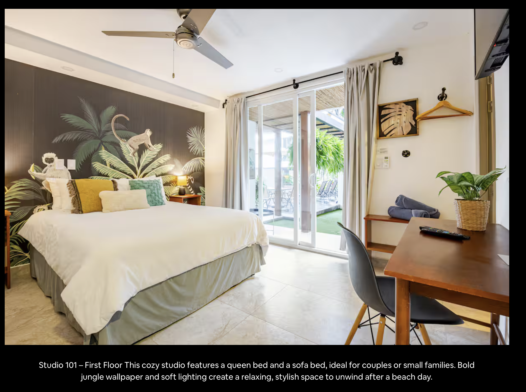 Cozy studio bedroom with a queen bed and a sofa bed, featuring a jungle-themed wall mural, pillows, and soft lighting, with sliding glass doors leading to an outdoor patio.