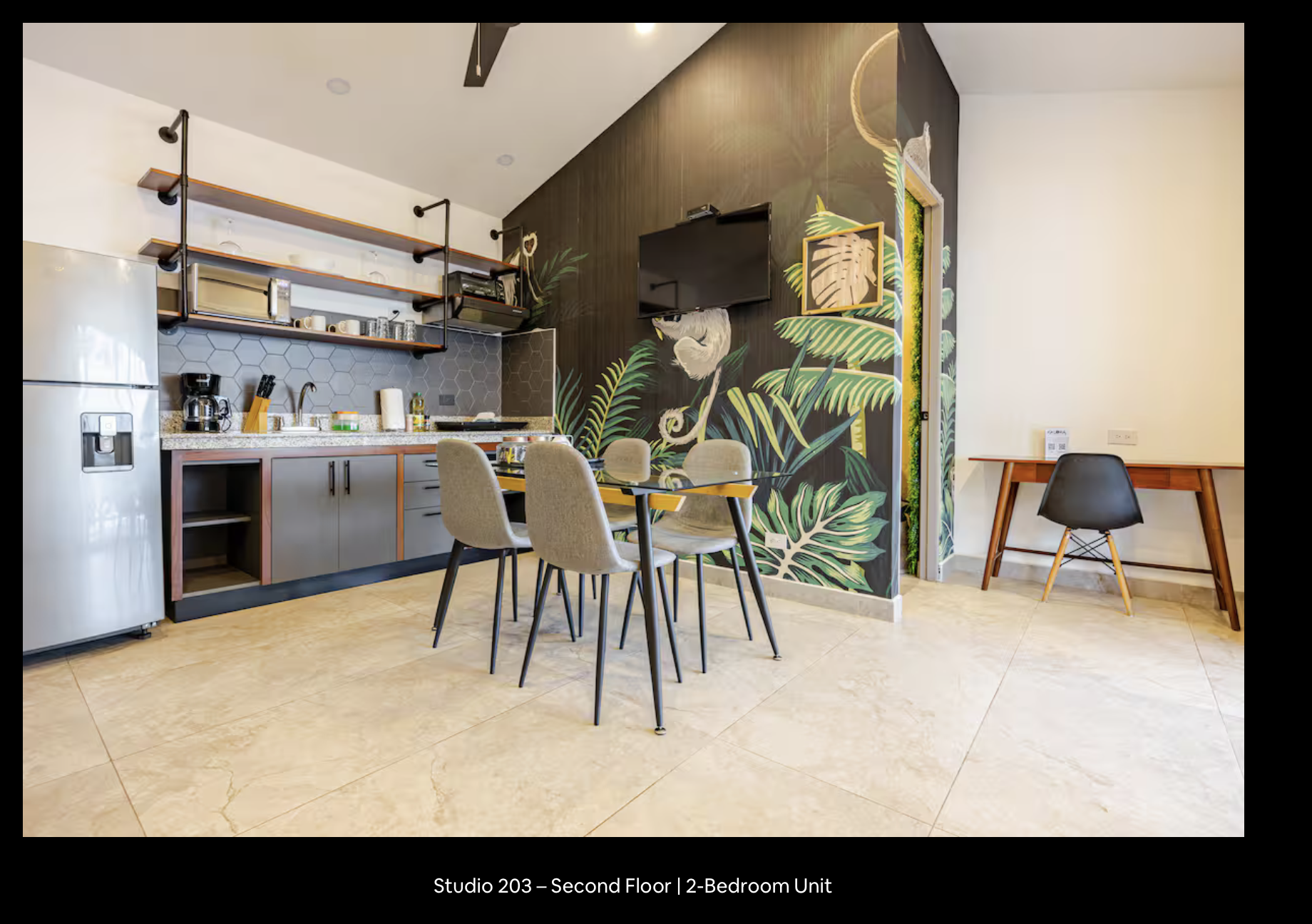 Interior of a modern kitchen and dining area with a tropical mural wall, featuring a refrigerator, open shelving, black hexagon backsplash, kitchen appliances, a dining table with six beige chairs, and a small workspace with a desk and chair.