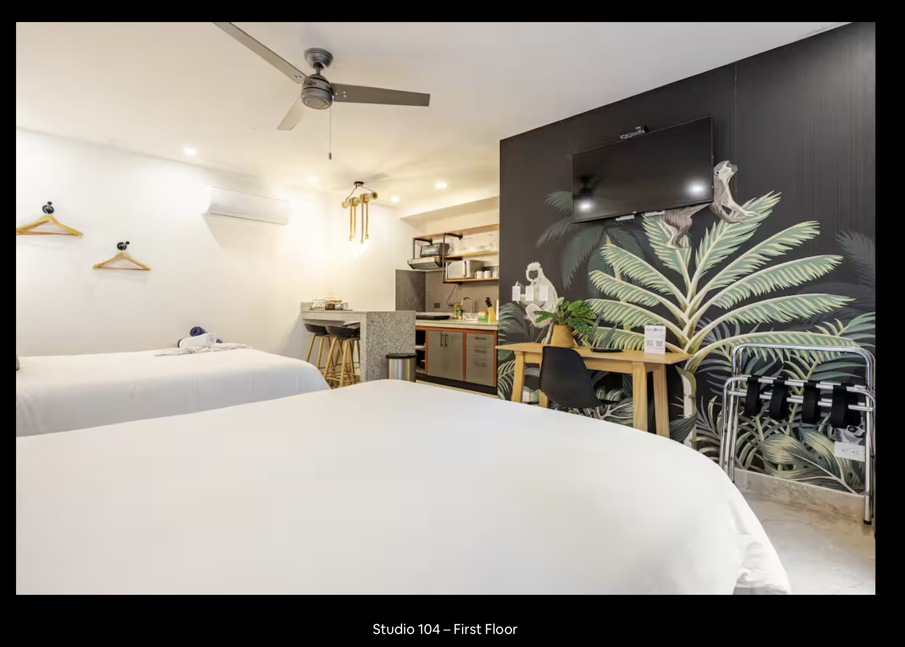 A hotel room with two white beds, a small kitchenette with bar stools, a wall-mounted TV, a wooden desk with a plant, black chairs, and a tropical leaf mural on the wall.