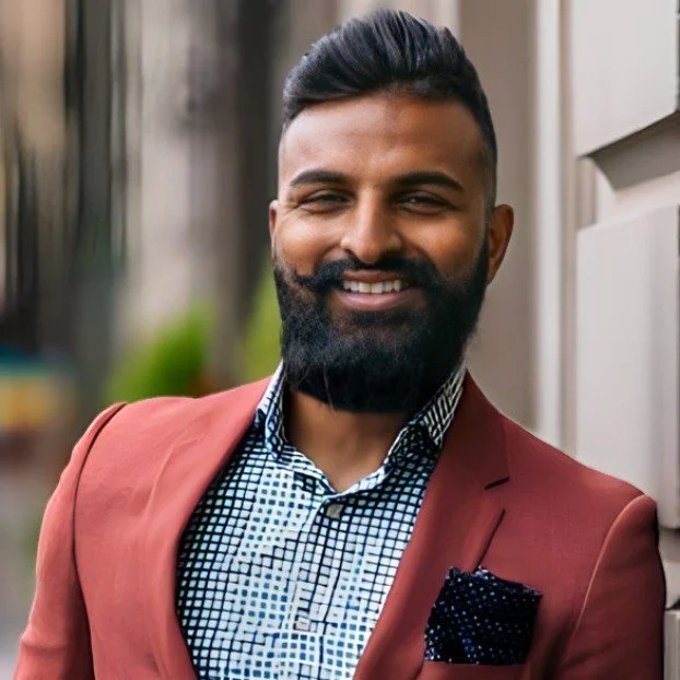 A man with a well-groomed beard and short hair, dressed in a checkered shirt and a reddish-brown blazer, smiling outdoors near a building with a blurred background.