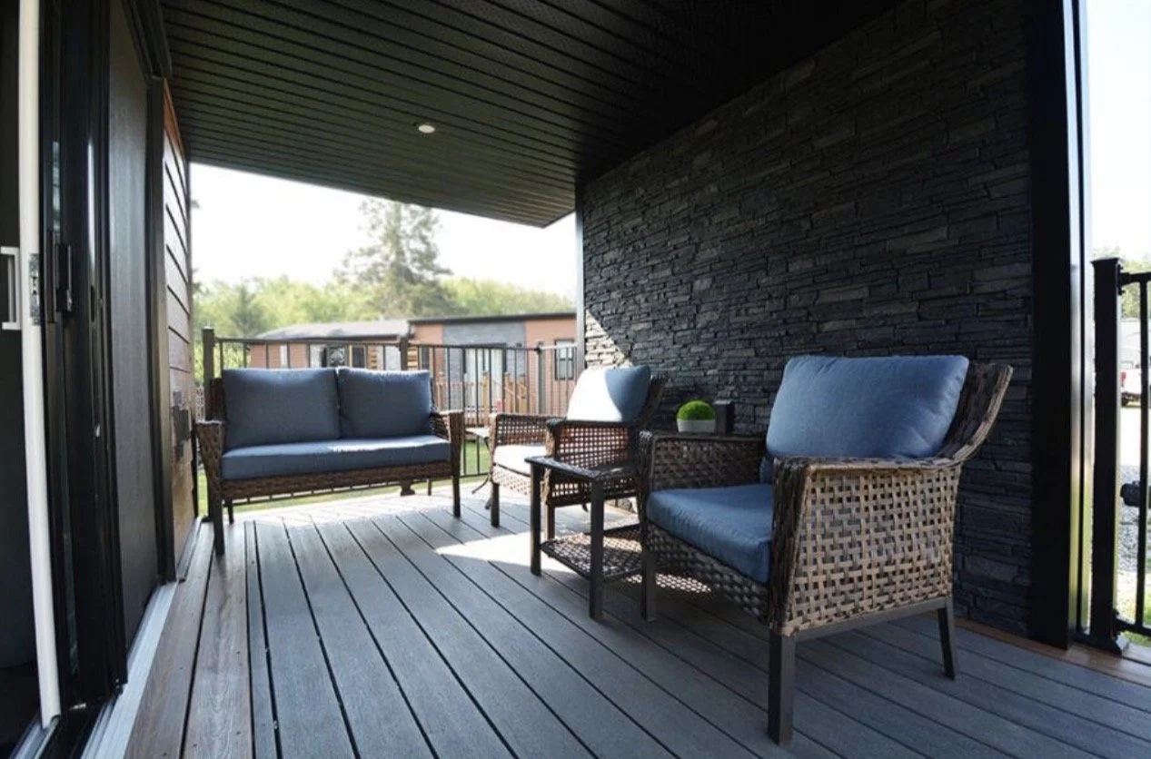Outdoor balcony with wicker furniture, including a sofa and two armchairs with blue cushions, a small side table with a plant, and a wooden railing overlooking trees and neighboring buildings.